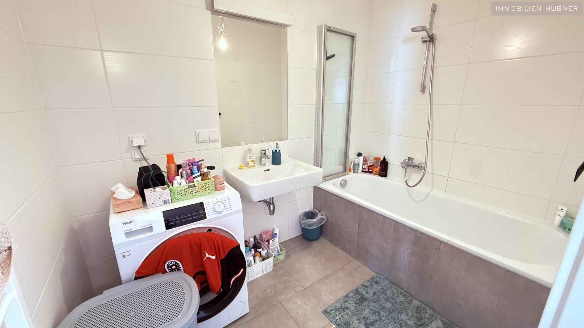 Badezimmer mit Badewanne, Waschbecken und Waschmaschine, hell gefliest.