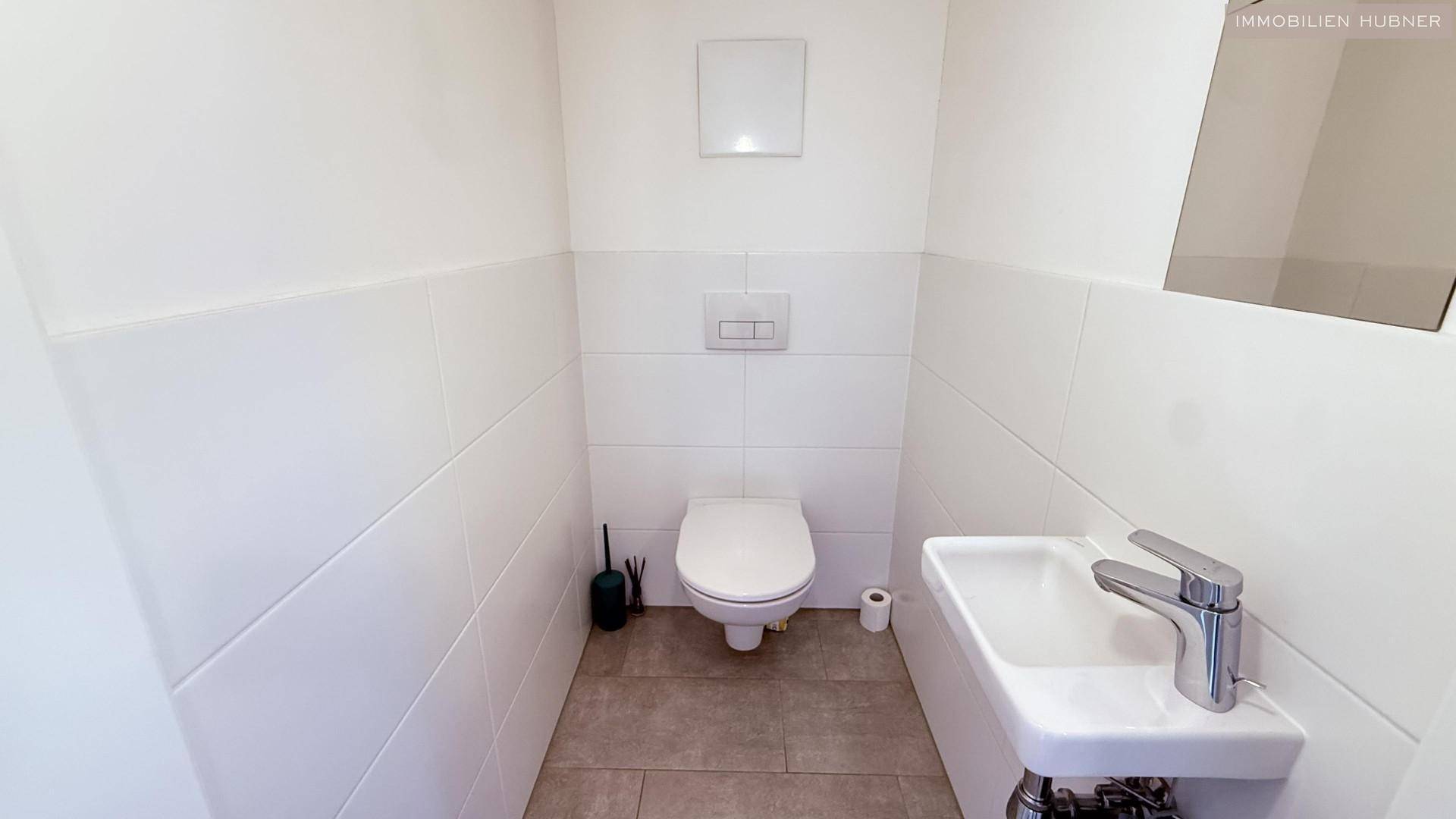 Separates WC mit Toilette und kleinem Handwaschbecken, weiß gefliest.