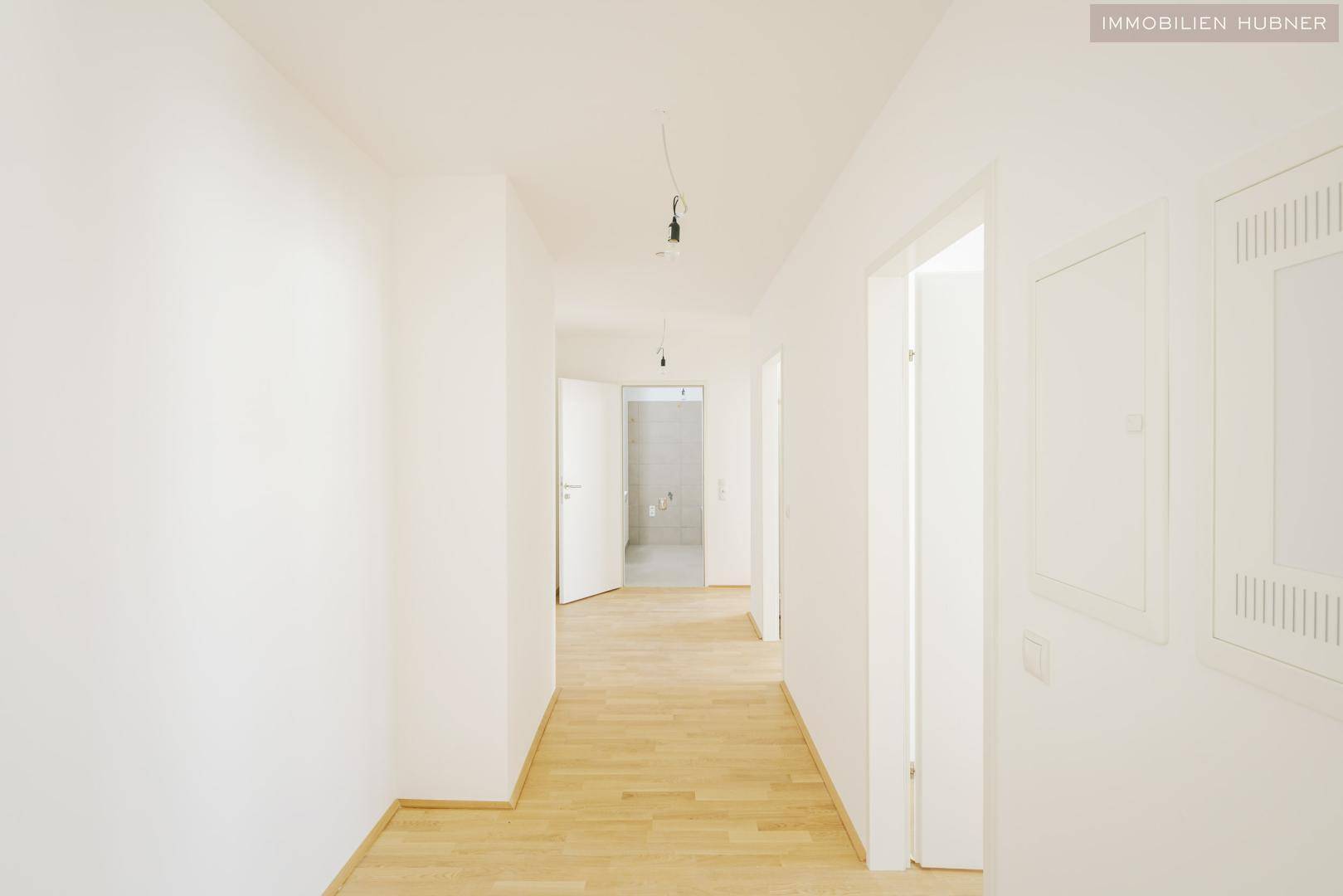 Langer, heller Flur mit Parkettboden und weißen Wänden, der einen Blick in ein Badezimmer ermöglicht.