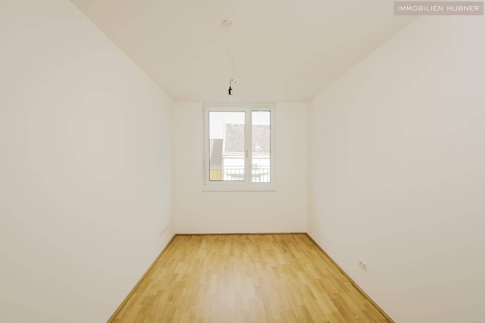 Leeres Schlafzimmer mit Parkettboden und einem Fenster, das viel Tageslicht hereinlässt.