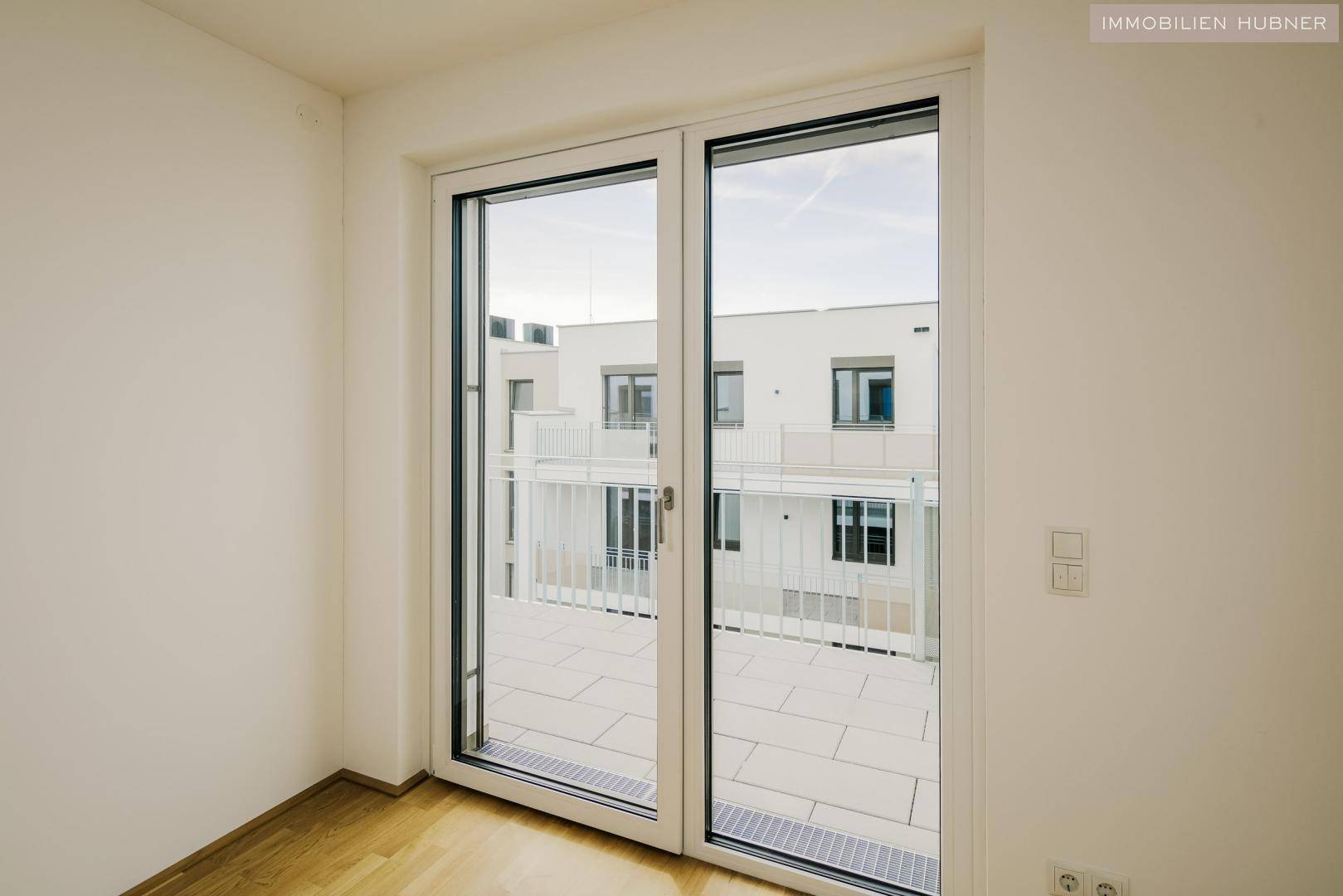 Großes Fenster mit Blick auf eine geflieste Terrasse und die moderne Architektur des Nachbargebäudes.