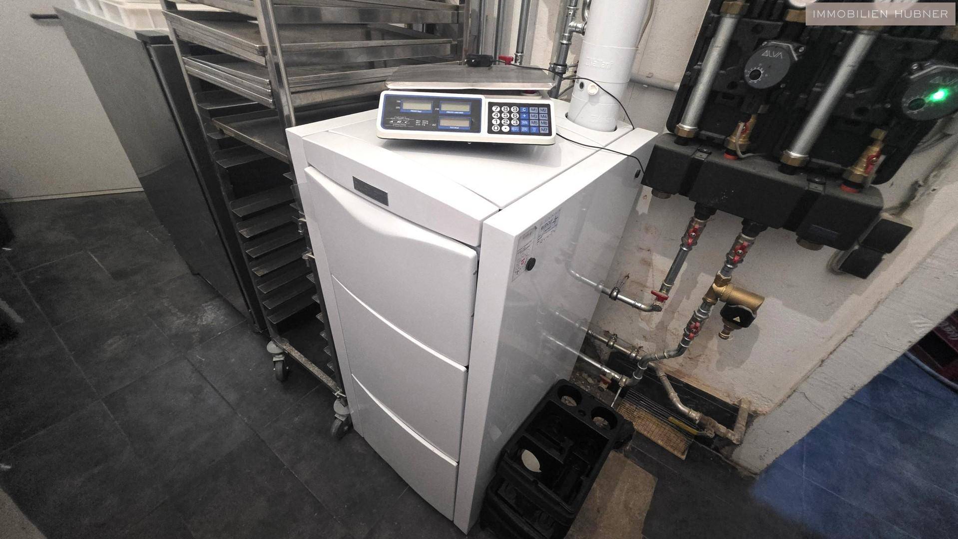 Technikraum mit Heizungsanlage, Waage und industriellen Regalen auf dunklem Boden.