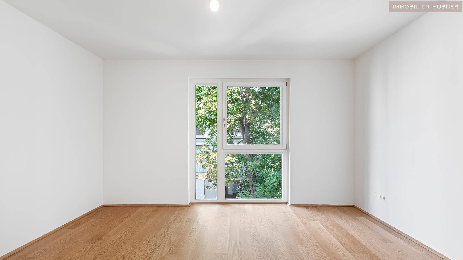 Helles Zimmer mit Holzboden und großem Fenster mit Blick ins Grüne.