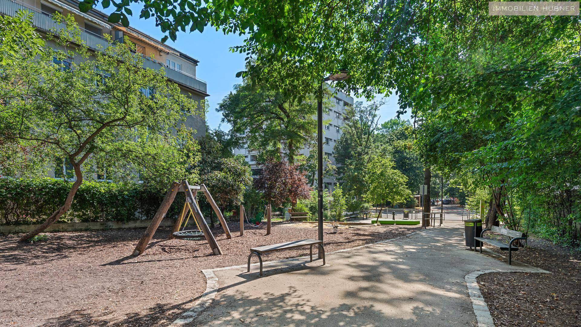 Grüner Spielplatz mit Schaukeln und Bänken, umgeben von Bäumen und Wohngebäuden.