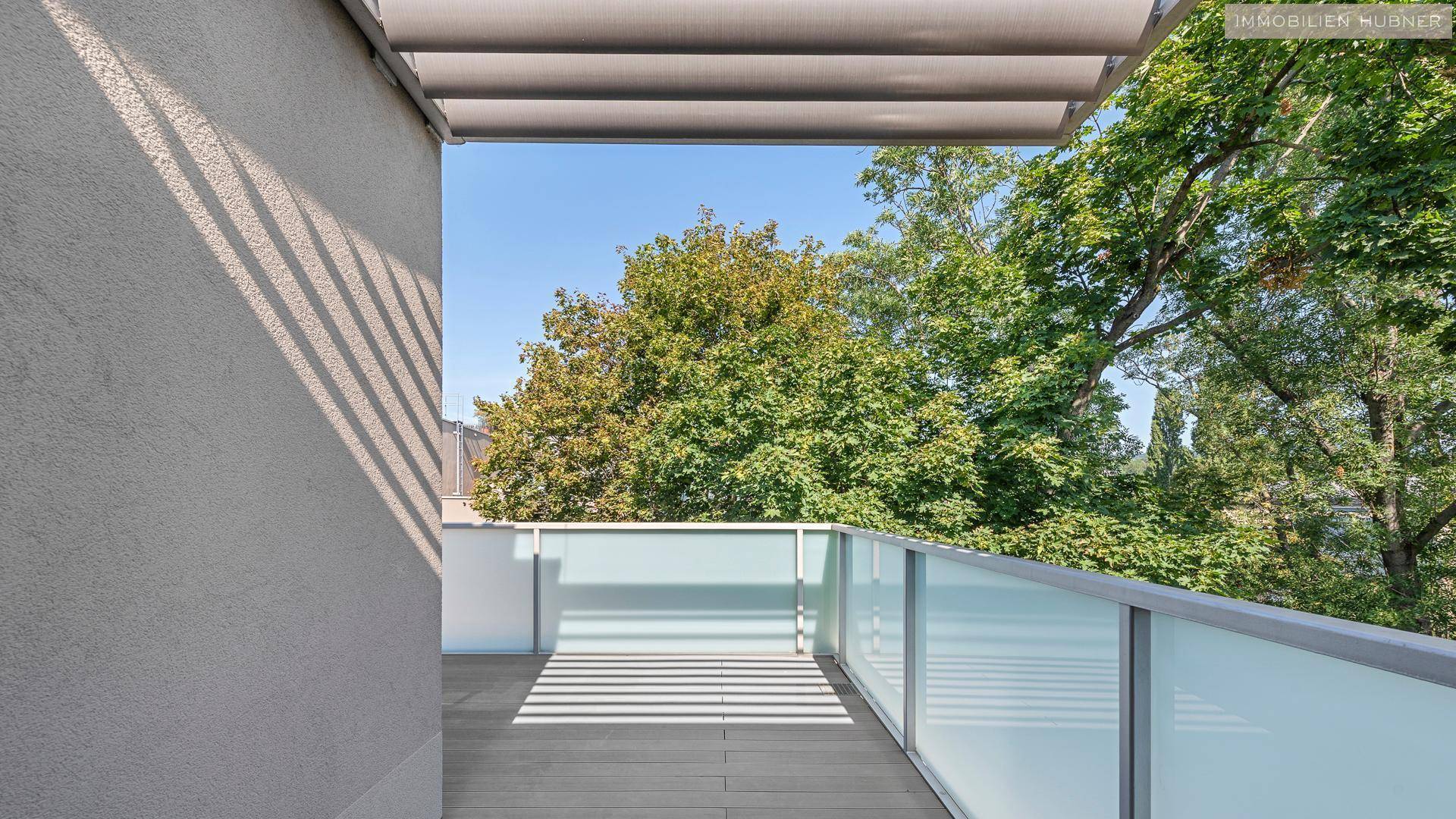 Sonniger Terrassenbereich mit Glasgeländer und Blick auf grüne Bäume.