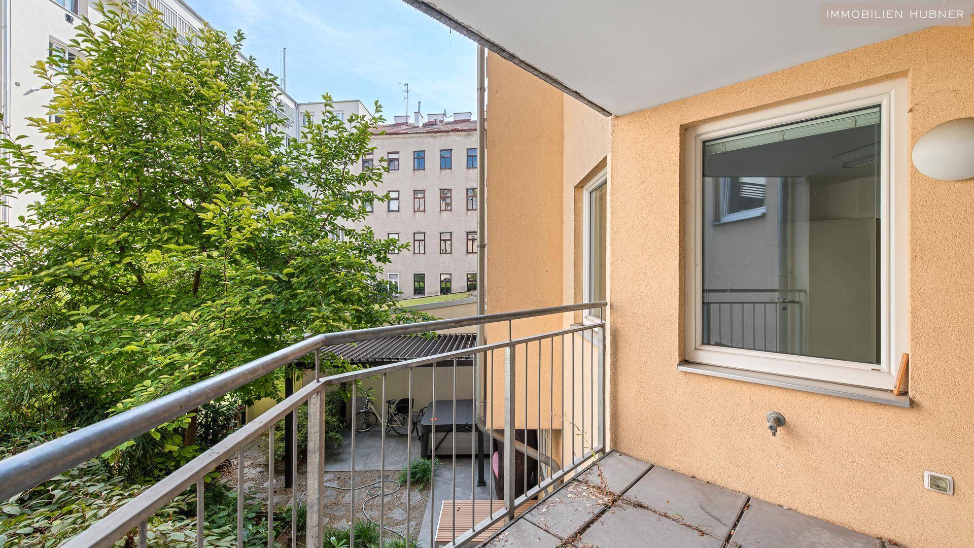 Kleiner Balkon mit Metallgeländer und Blick auf einen begrünten Innenhof und umliegende Gebäude.