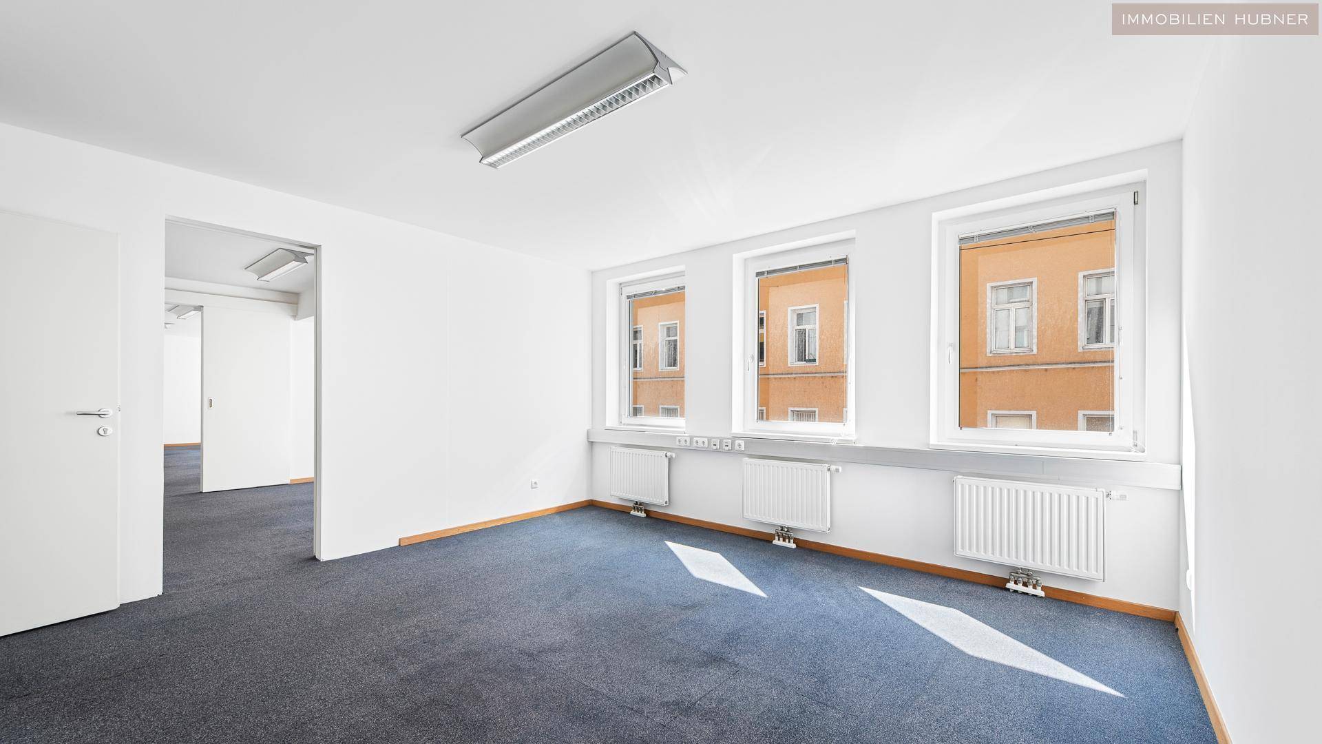 Heller Büroraum mit Teppichboden und drei Fenstern, die einen Blick auf die gegenüberliegenden Gebäude bieten.
