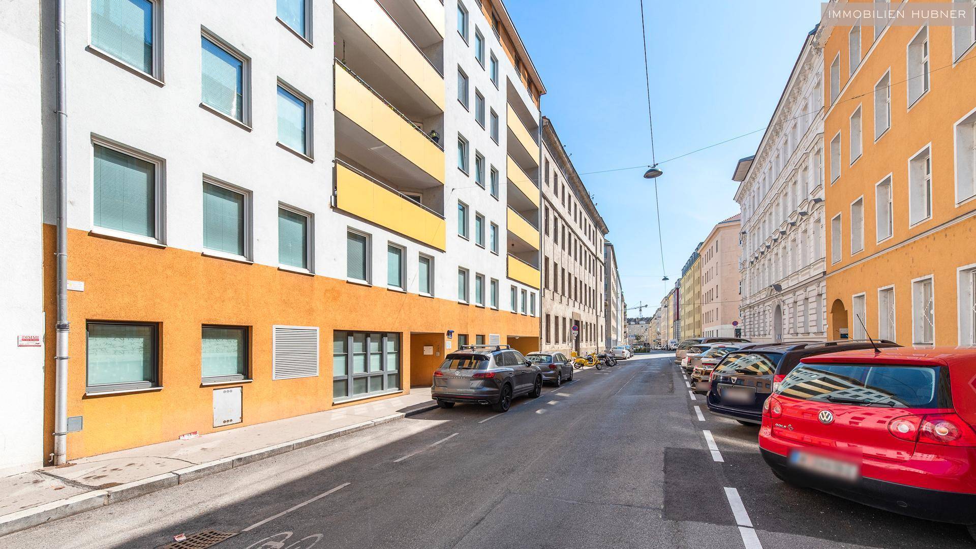 Straßenansicht mit mehrstöckigen Gebäuden und parkenden Autos unter blauem Himmel.