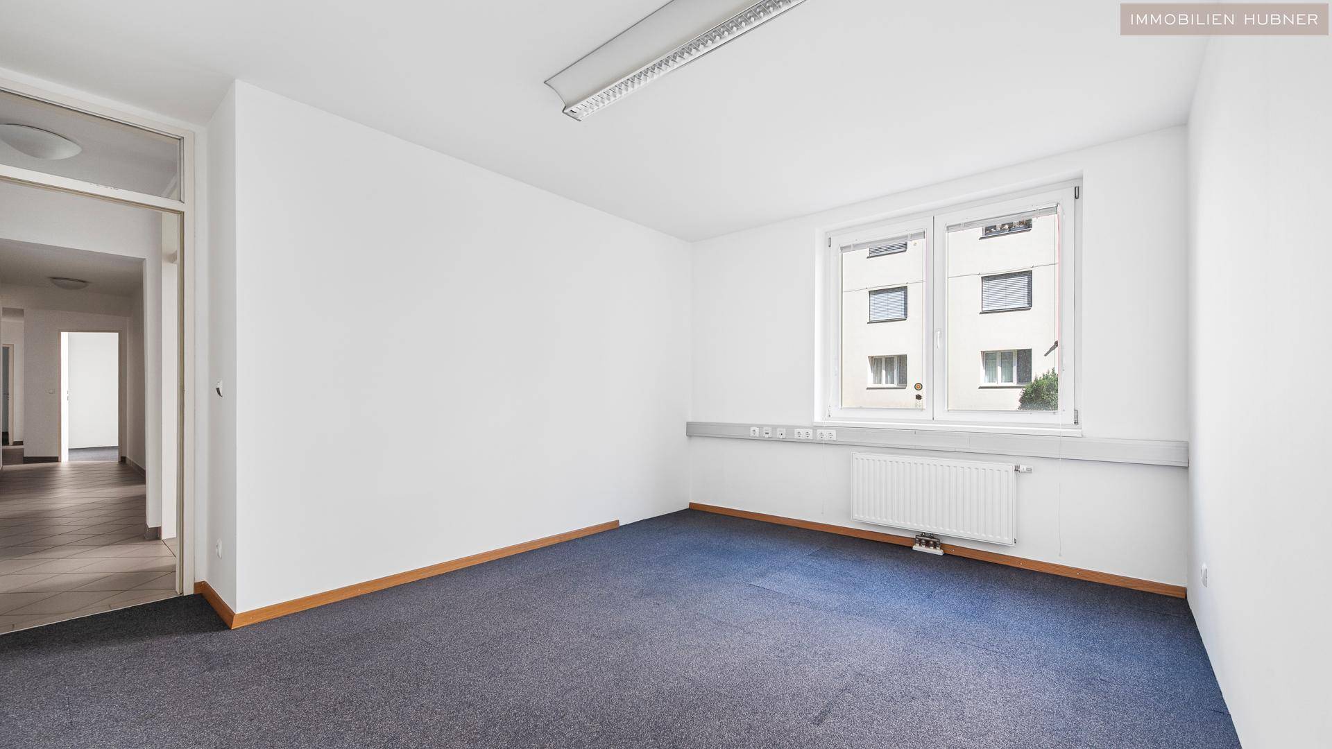Heller Büroraum mit Teppichboden und einem großen Fenster, das den Blick auf die Umgebung freigibt.