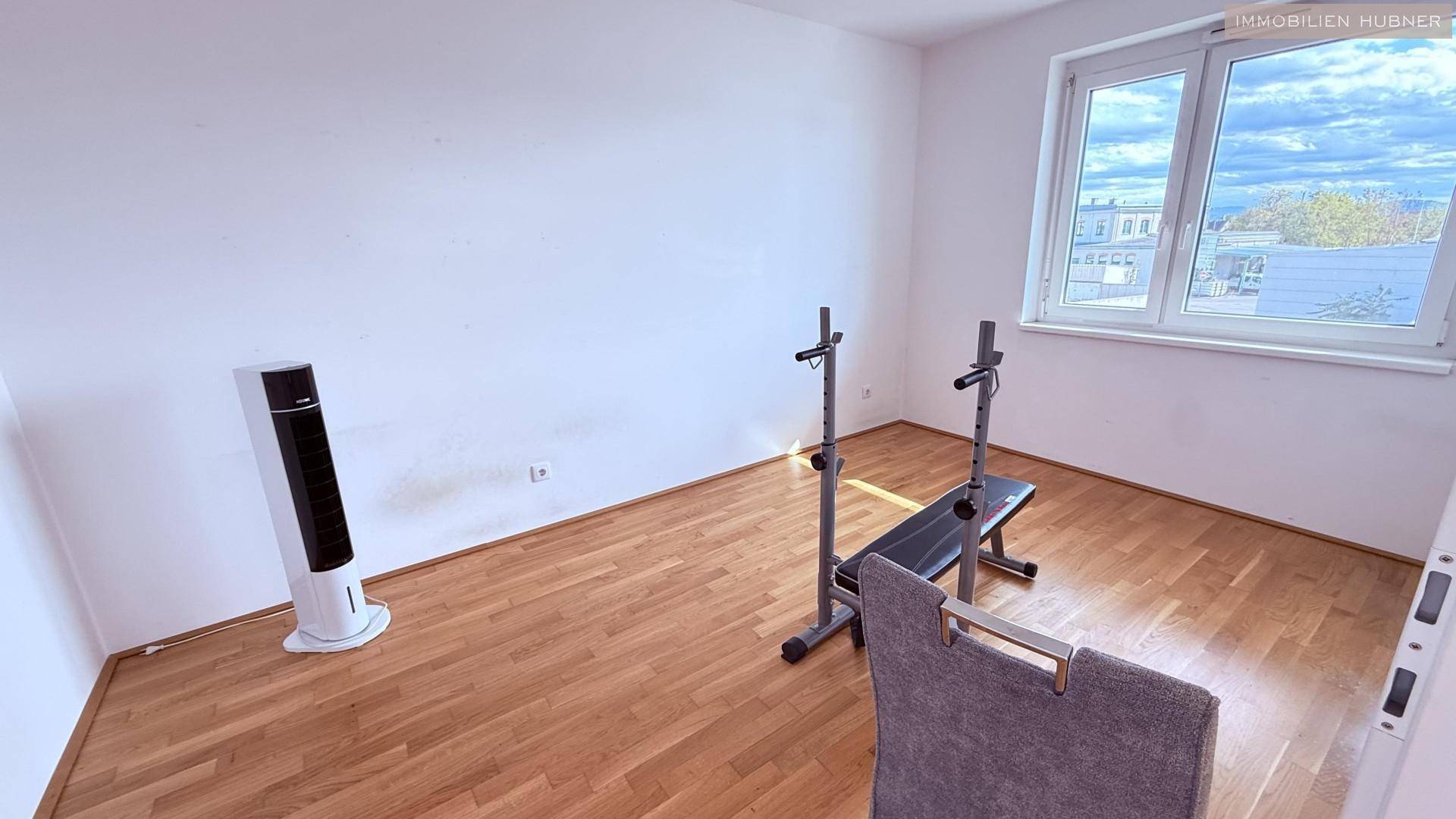 Helles Zimmer mit Parkettboden und Fenster, ausgestattet mit einem Trainingsgerät.
