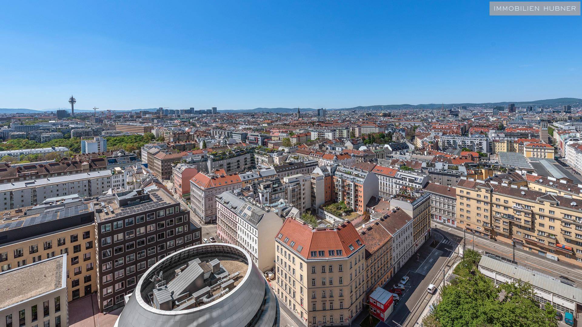 Weitläufiger Blick über die Stadt mit dichter Bebauung und einem markanten Turm am Horizont.