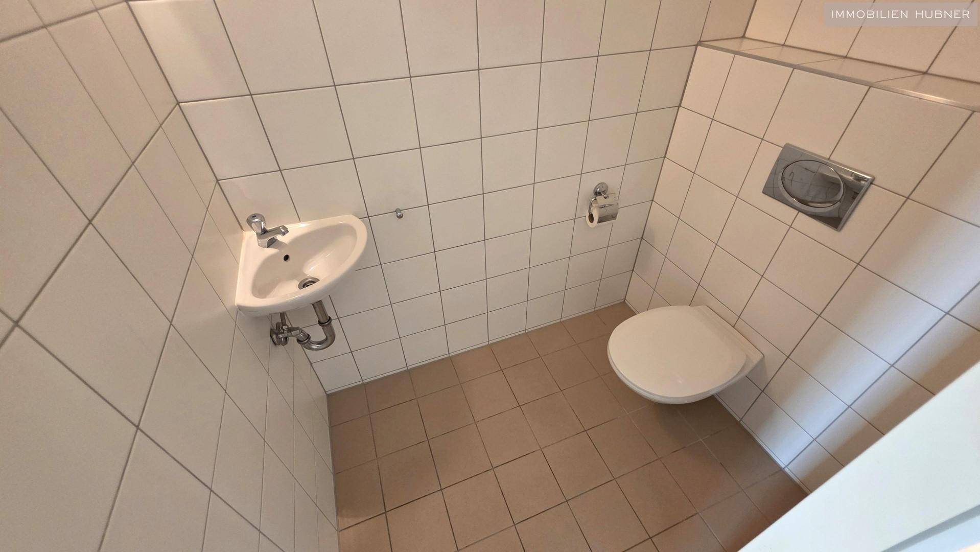 Separate Toilette mit Wand-WC, kleinem Waschbecken und hellen Fliesen.