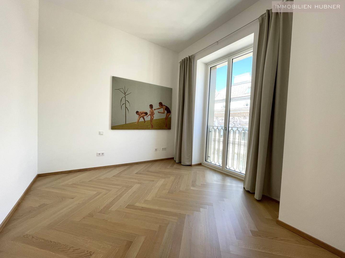 Geräumiges Zimmer mit Fischgrätparkett, großen Fenstern und einem französischen Balkon.
