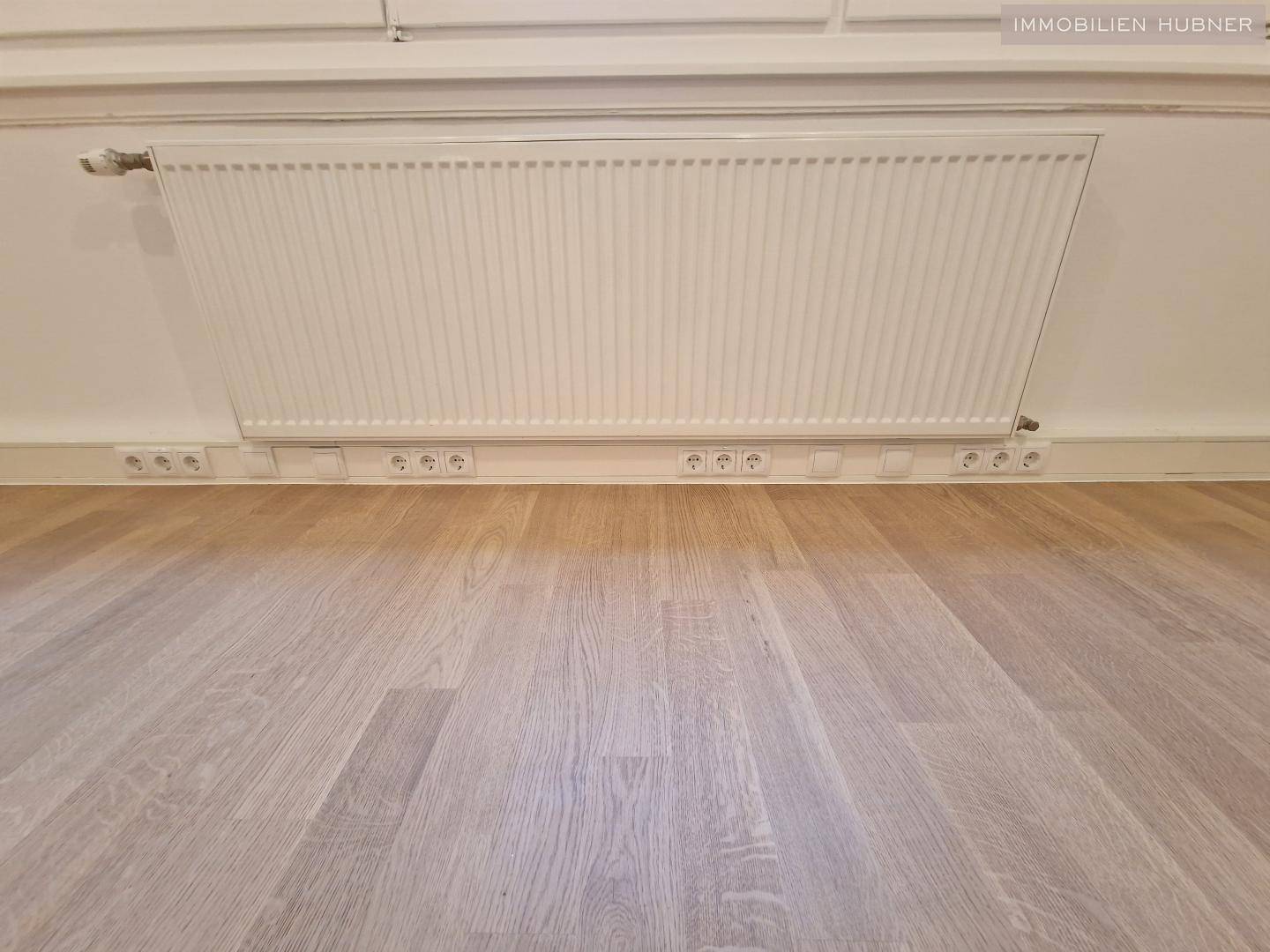Detailaufnahme eines Heizkörpers und einer Reihe von Steckdosen entlang der Fußleiste.