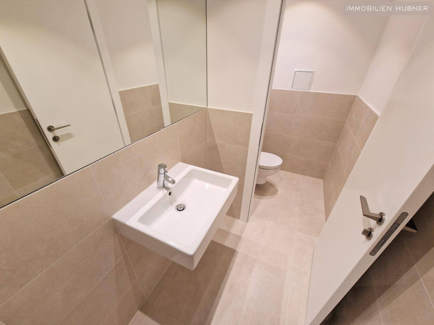 Modernes Gäste-WC mit schwebendem Waschbecken, großem Spiegel und beigefarbenen Fliesen.