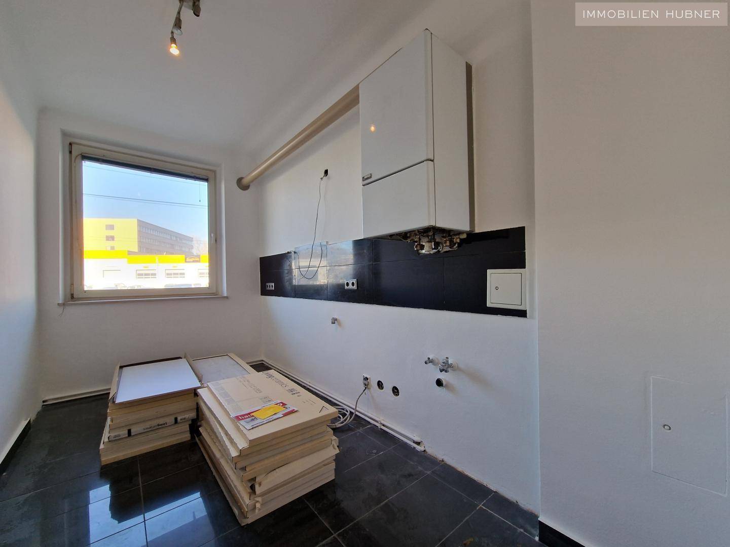 Leerer Küchenraum mit dunklen Fliesen, Fenster und Anschlüssen für eine neue Einbauküche.
