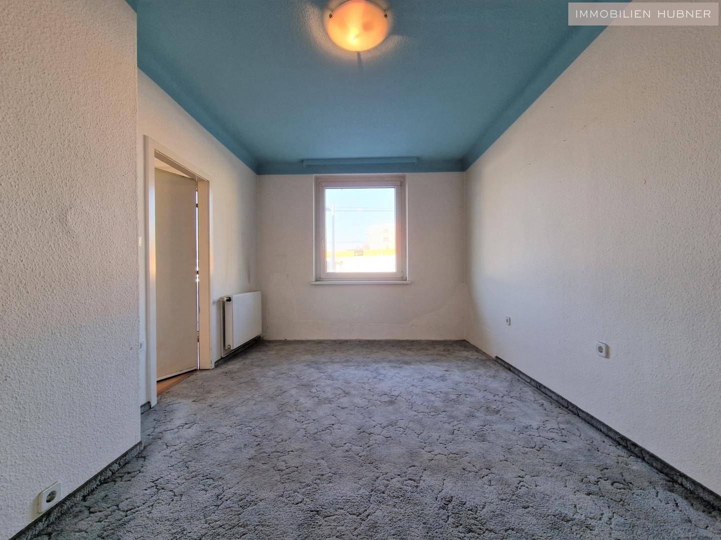 Leeres Zimmer mit blau gestrichener Decke, Fenster und Teppichboden.