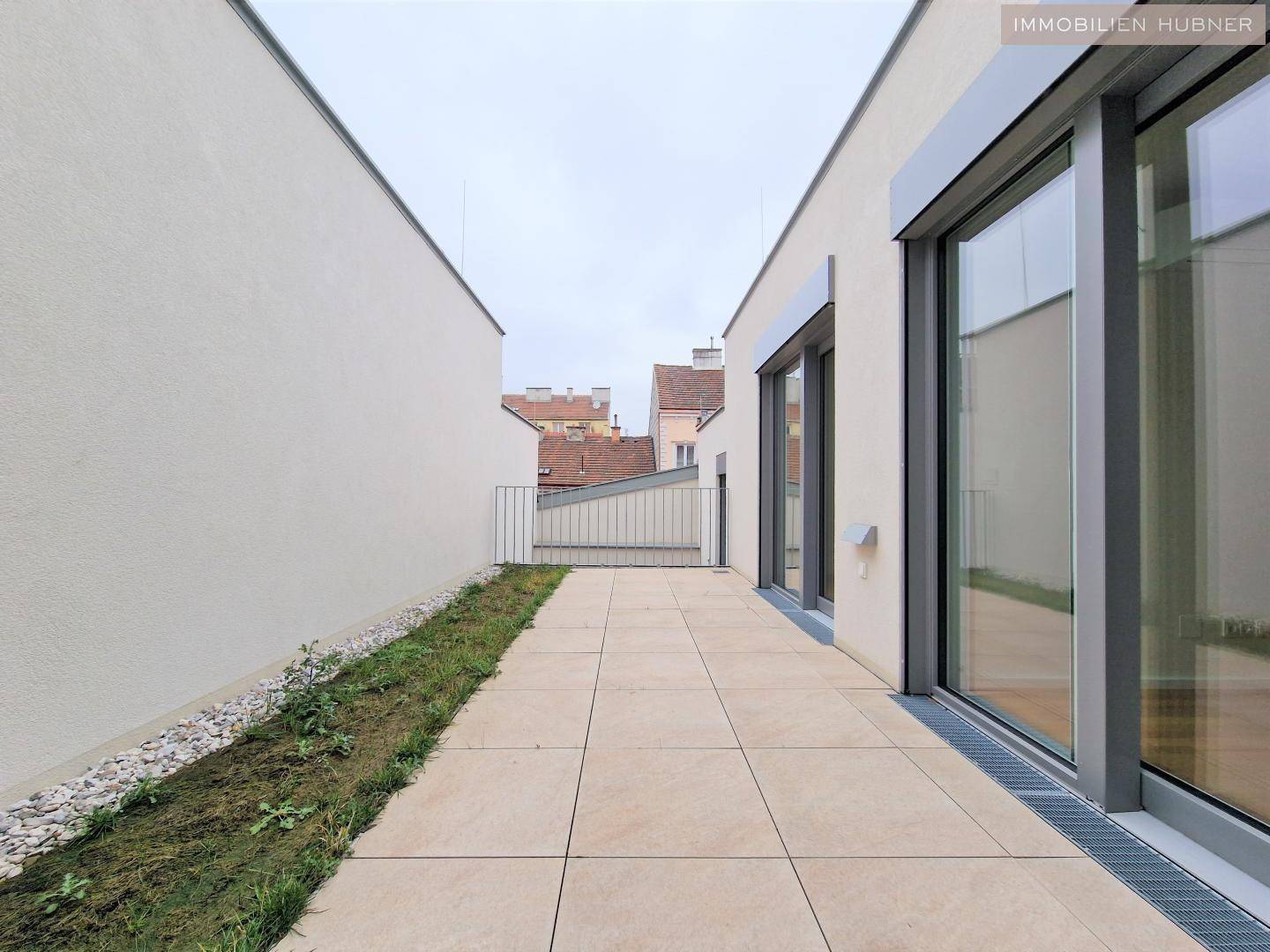 Lange, geflieste Terrasse mit kleiner Grünfläche und Blick auf die umliegenden Gebäude.