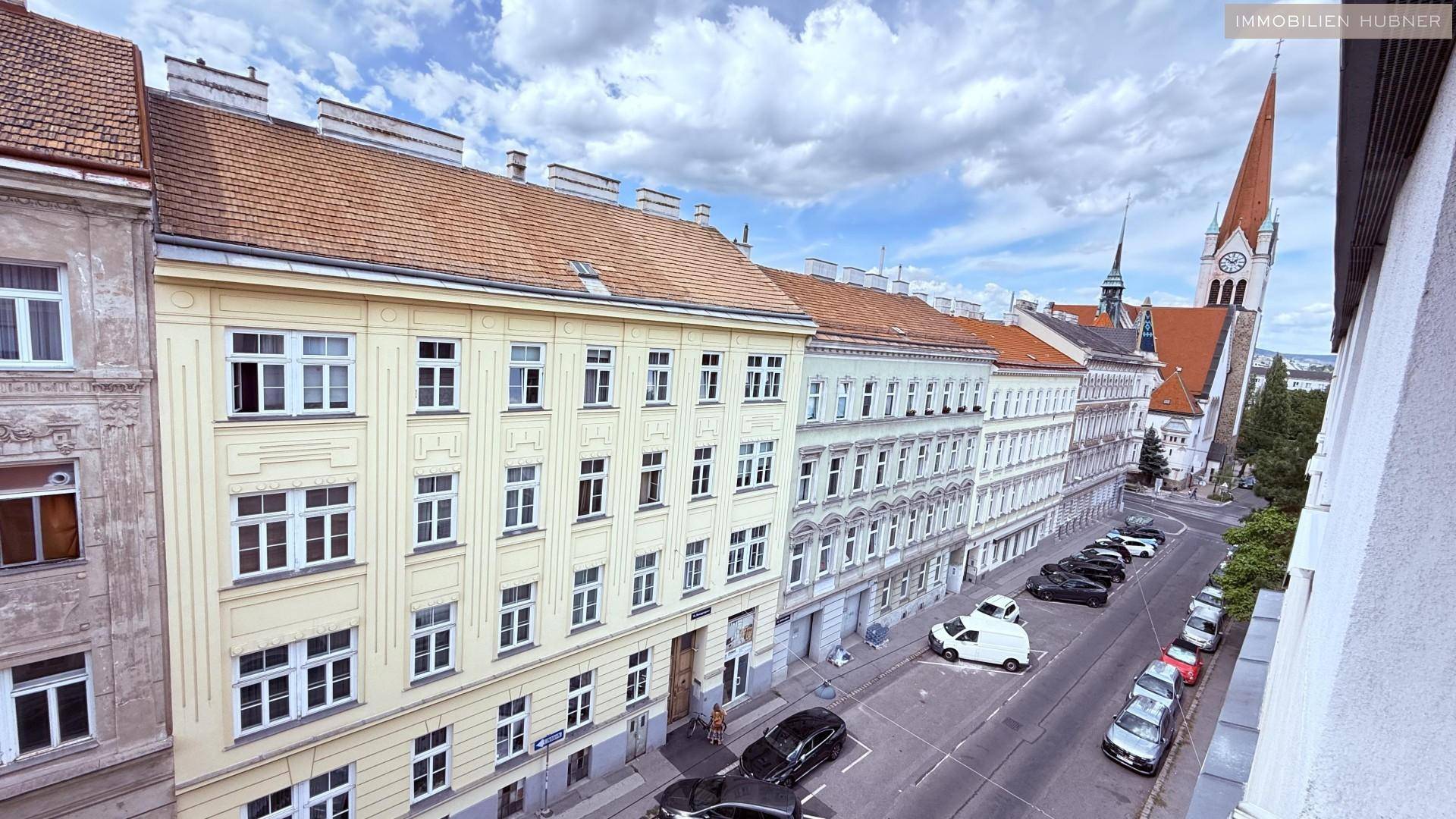 Blick von oben auf eine belebte Straße mit parkenden Autos und einem Kirchturm im Hintergrund.