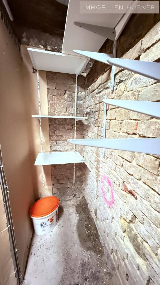 Ecke eines Kellerraums mit Ziegelwänden, Betonboden und praktischen Wandregalen unter einer Treppe.