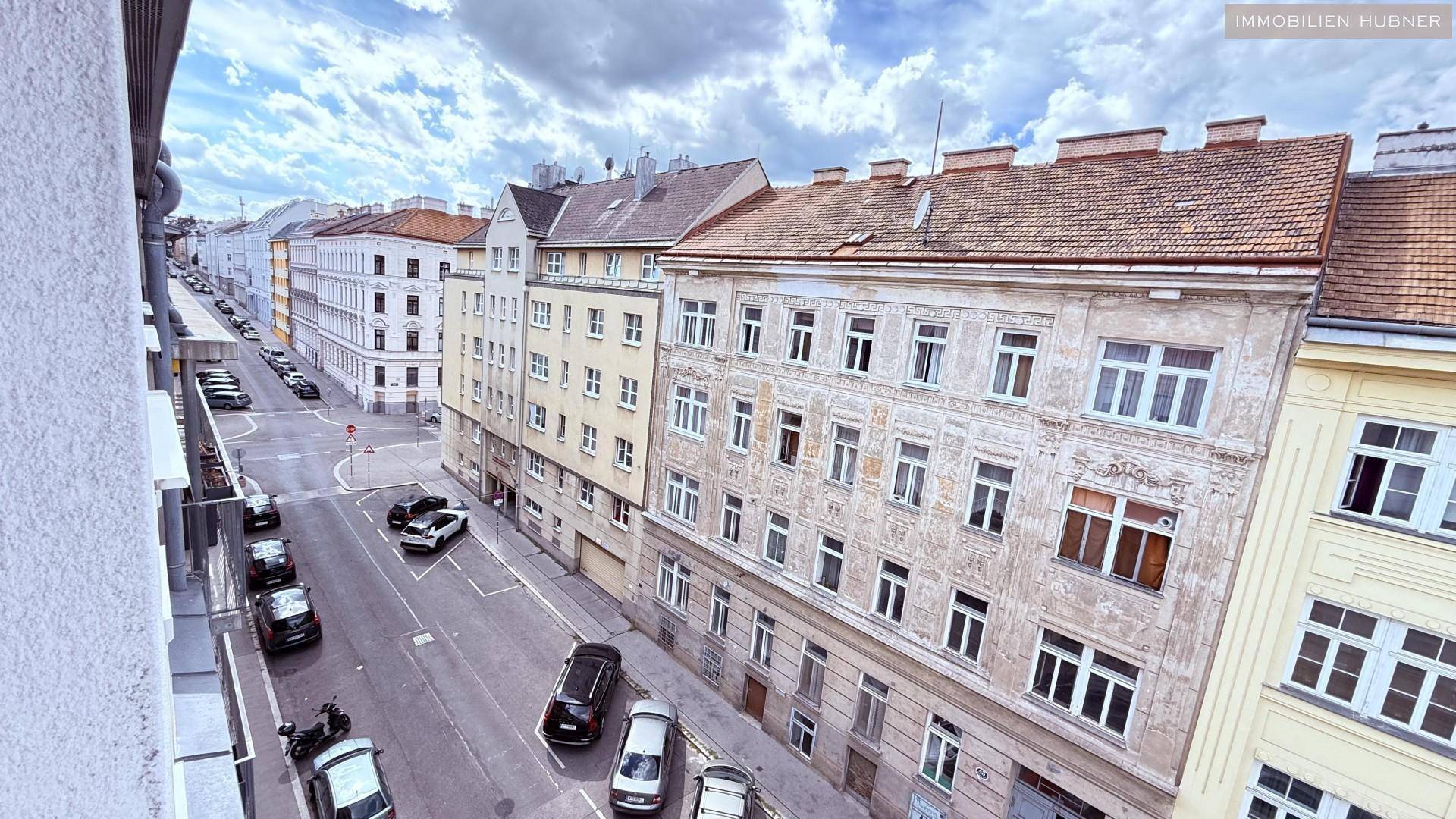 Panoramablick auf eine historische Straße mit Altbauten und geparkten Fahrzeugen unter bewölktem Himmel.