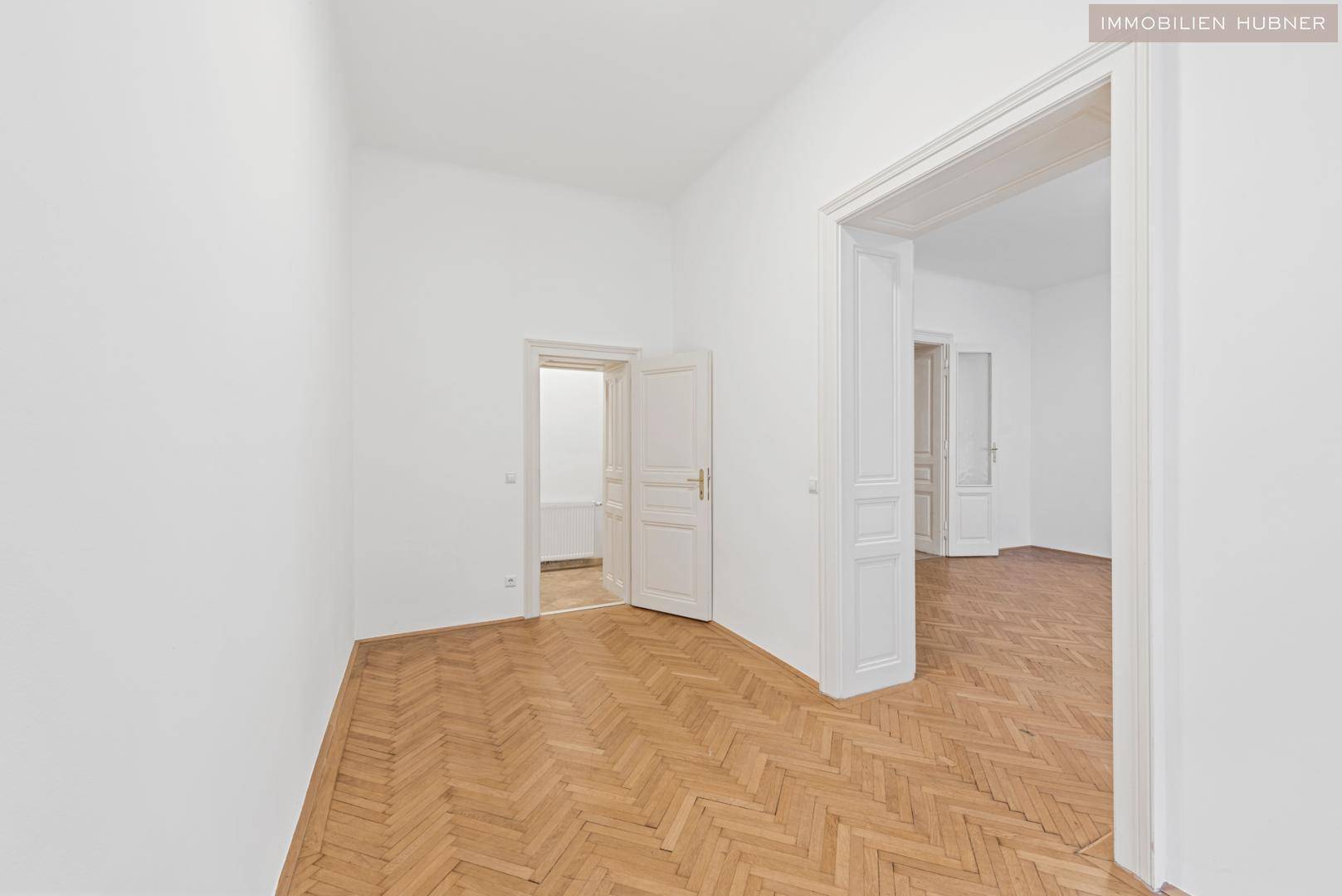 Helles Zimmer mit Fischgrätparkett und Zugang zu weiteren Räumen sowie einem Badezimmer.