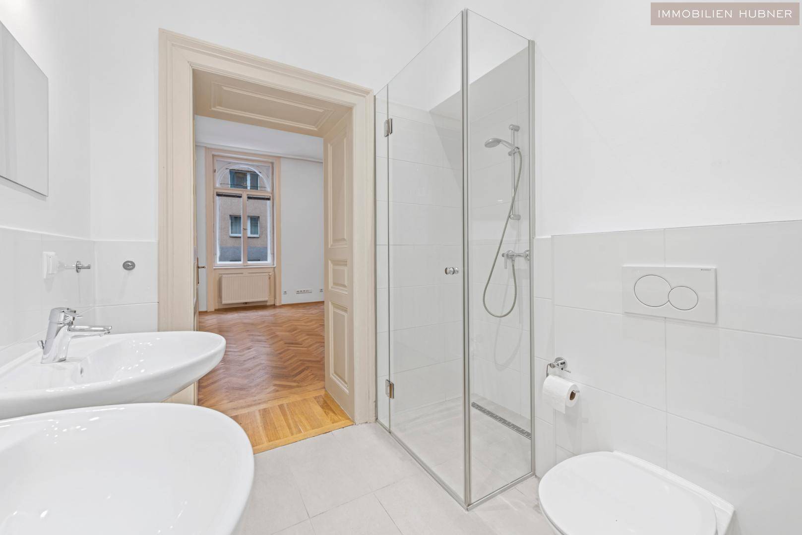 Helles Badezimmer mit Doppelwaschbecken, Duschkabine und Zugang zu einem Zimmer mit Parkettboden.