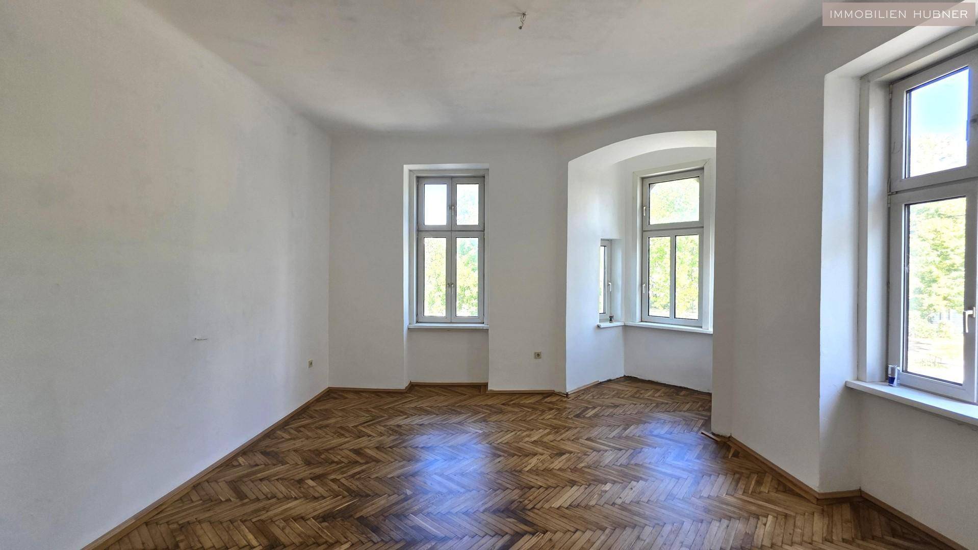 Geräumiges Zimmer mit Fischgrätparkett und vielen Fenstern, die viel Tageslicht hereinlassen.