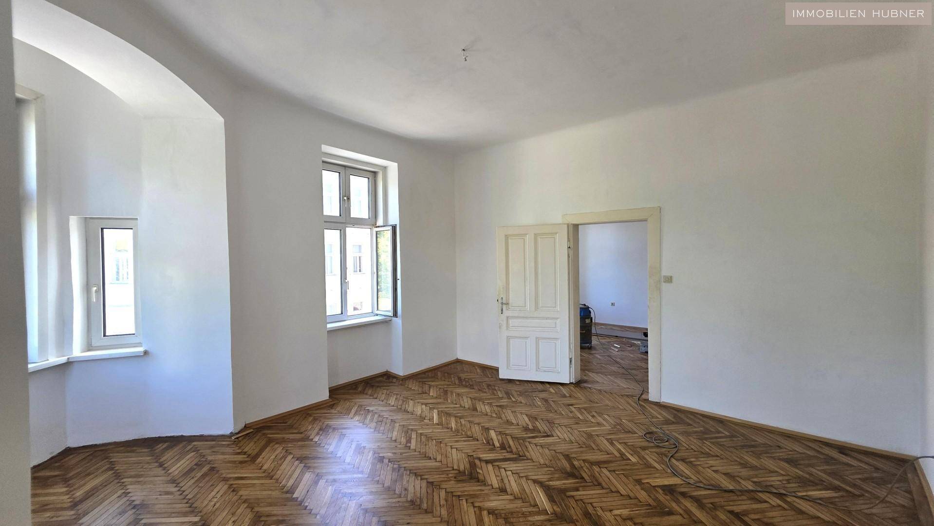 Lichtdurchflutetes Zimmer mit klassischem Fischgrätparkett und Zugang zu einem weiteren Raum.