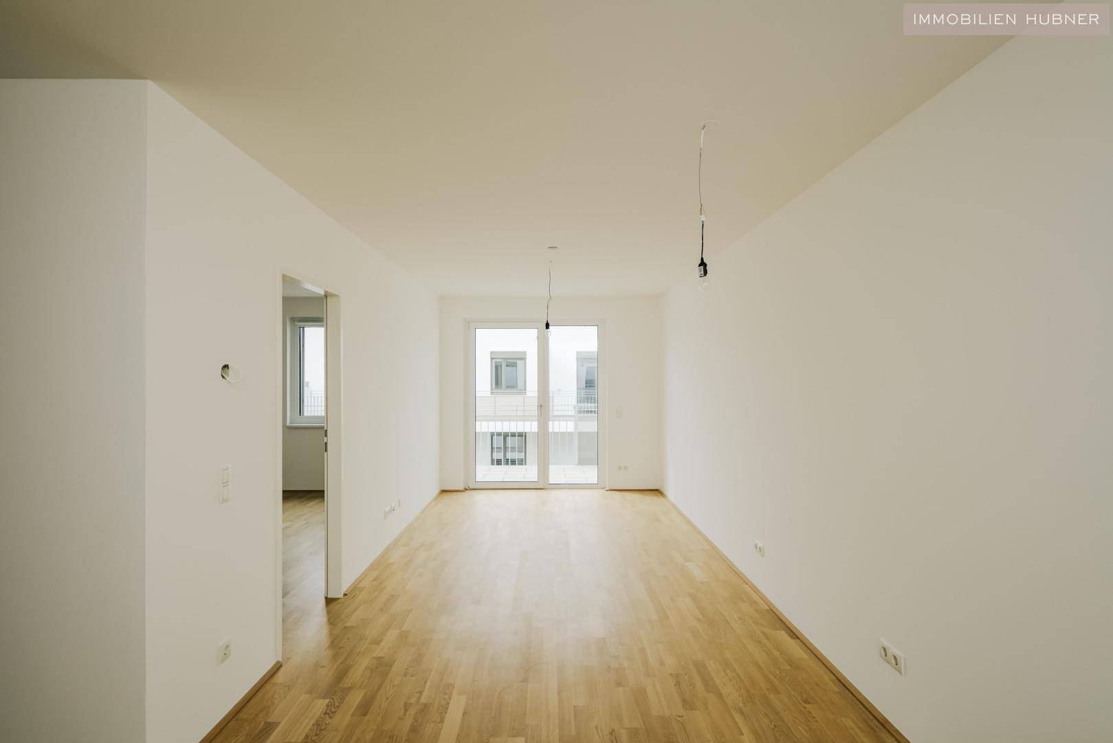 Helles, leeres Wohnzimmer mit Parkettboden und Zugang zu einem Balkon.