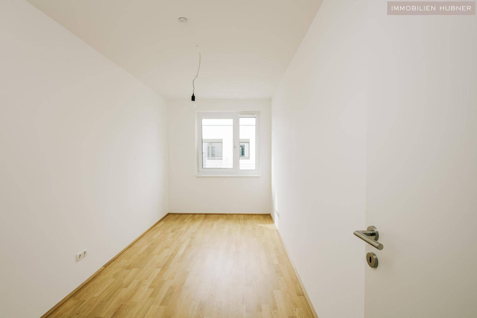 Helles Schlafzimmer mit Fenster, Parkettboden und weiß gestrichenen Wänden.