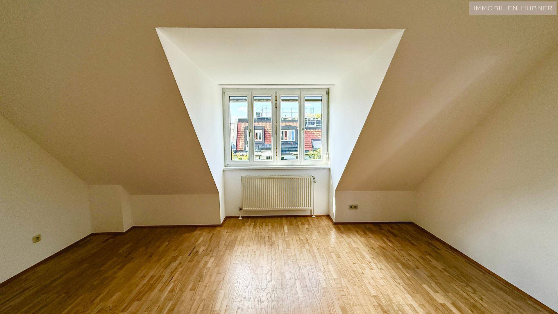 Helles Zimmer im Dachgeschoss mit Holzboden und großem Fenster, das viel Licht hereinlässt.
