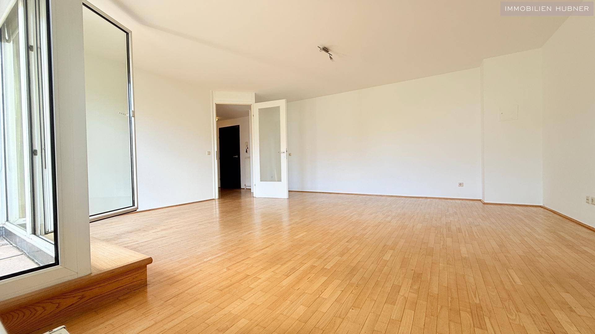Weitläufiges Wohnzimmer mit Parkettboden und großen Fenstern, die viel Tageslicht hereinlassen.