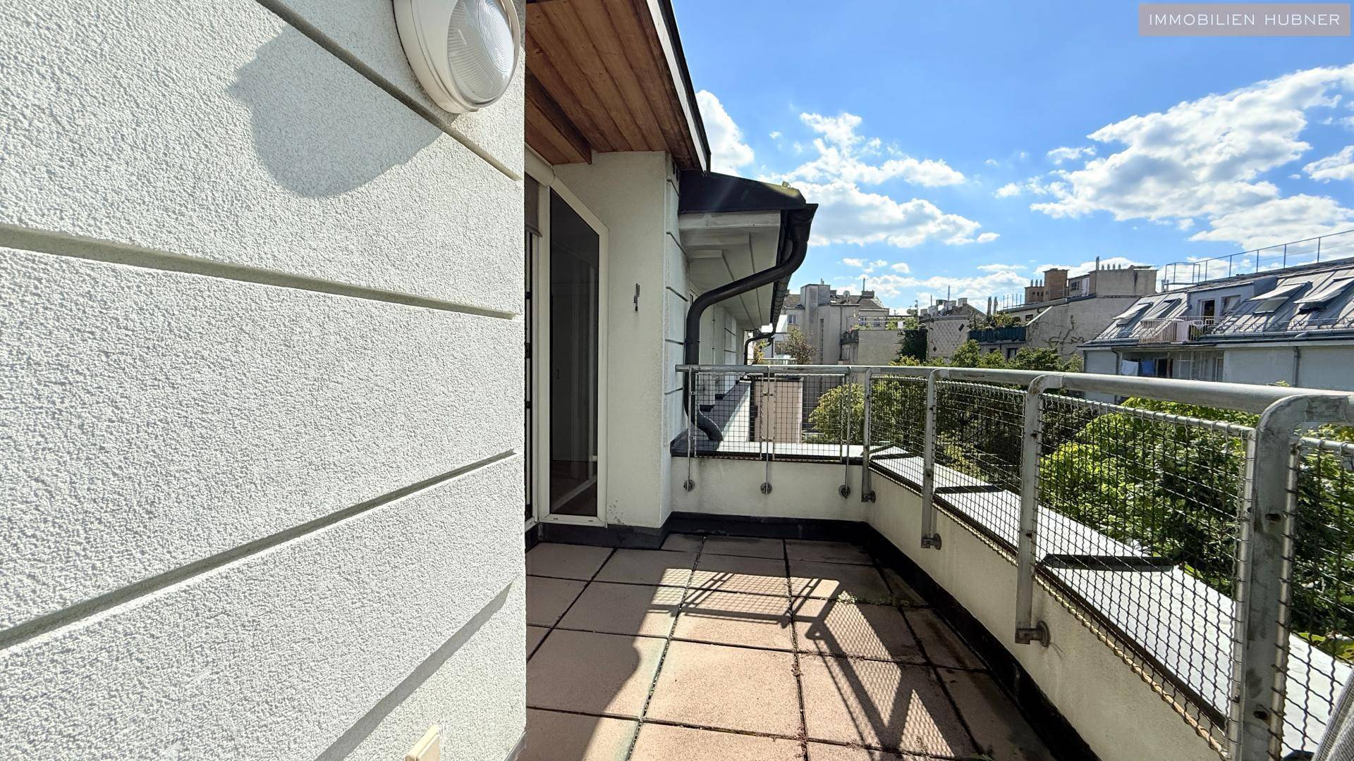 Langer Balkon mit Fliesenboden und Blick auf die umliegenden Gebäude und Bäume.