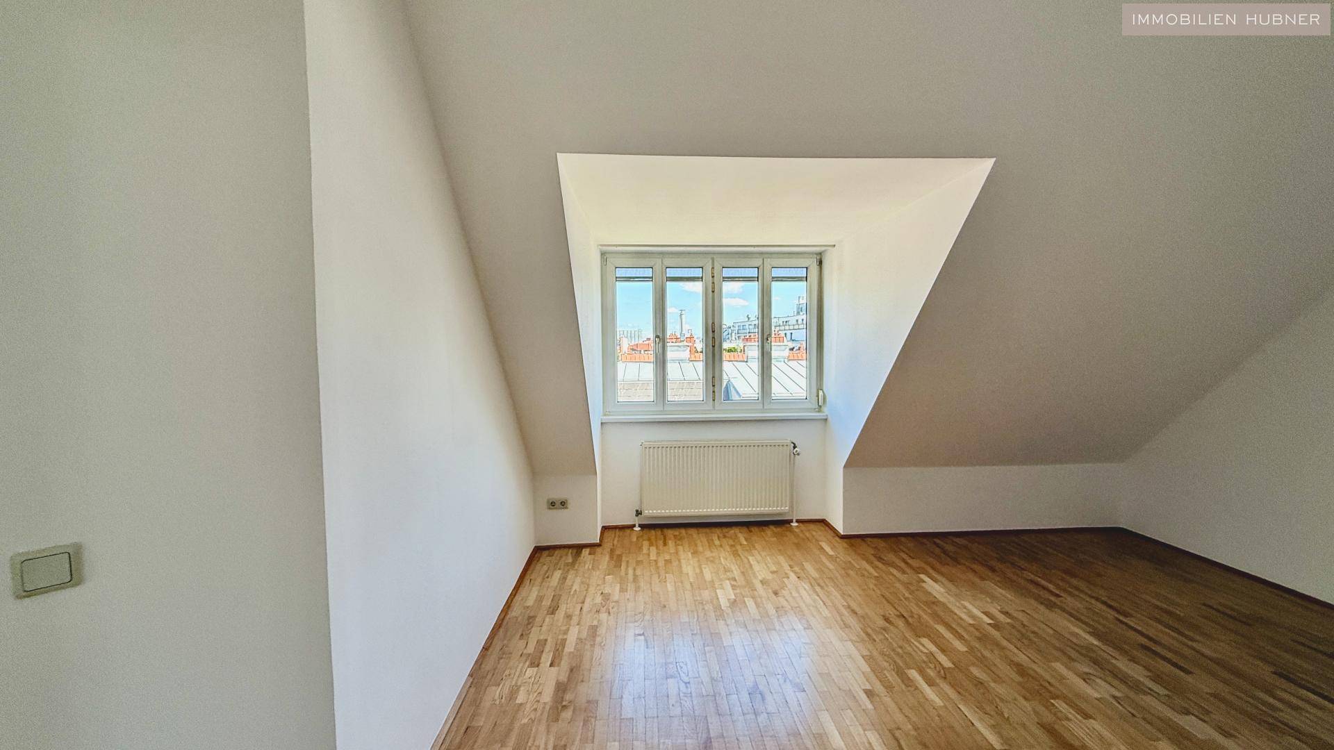 Geräumiges Zimmer im Dachgeschoss mit Holzboden und großem Fenster, ideal als Schlaf- oder Arbeitszimmer.