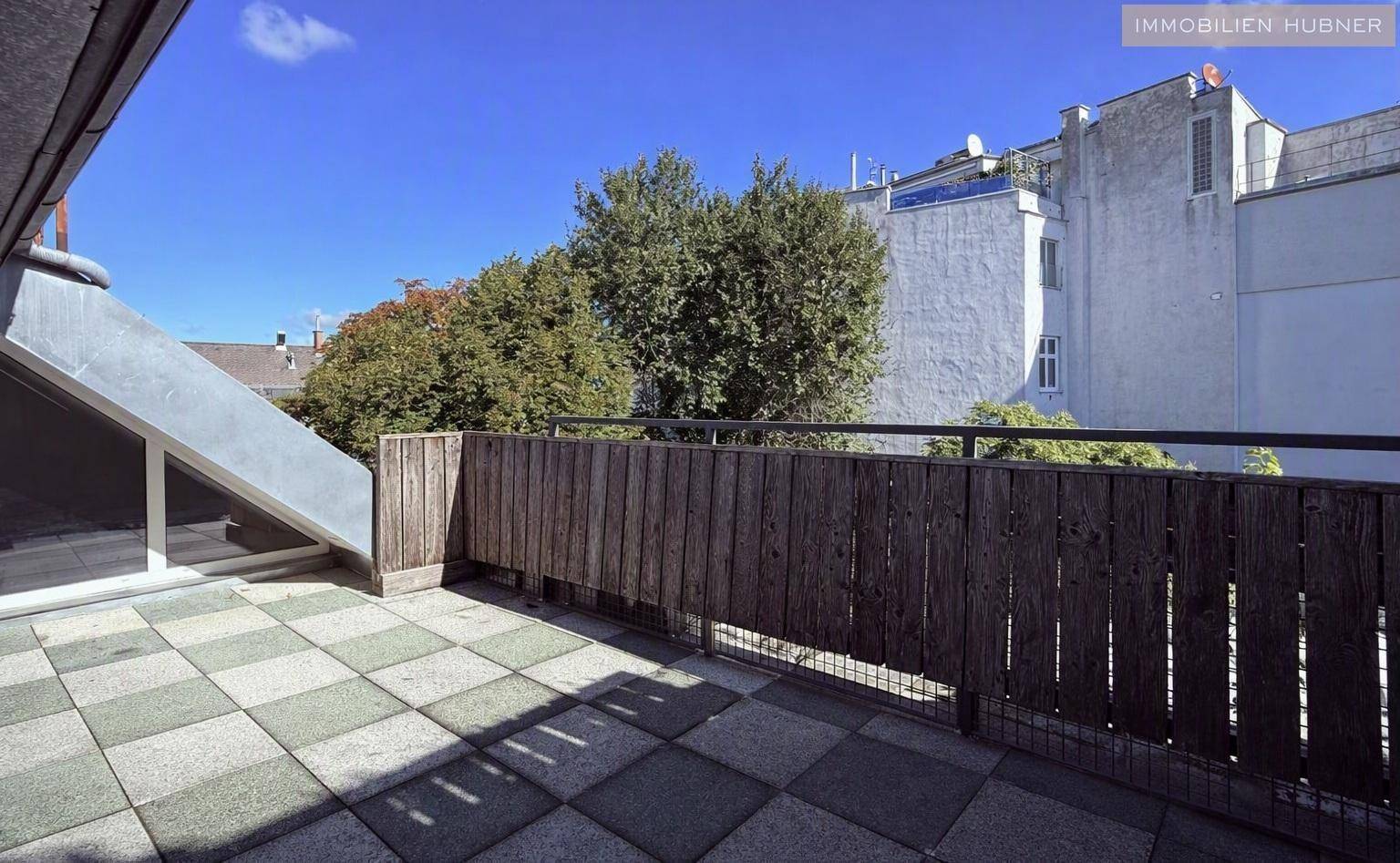 Geräumige Dachterrasse mit karierten Fliesen und Holzzaun, umgeben von Stadtgebäuden und Bäumen.