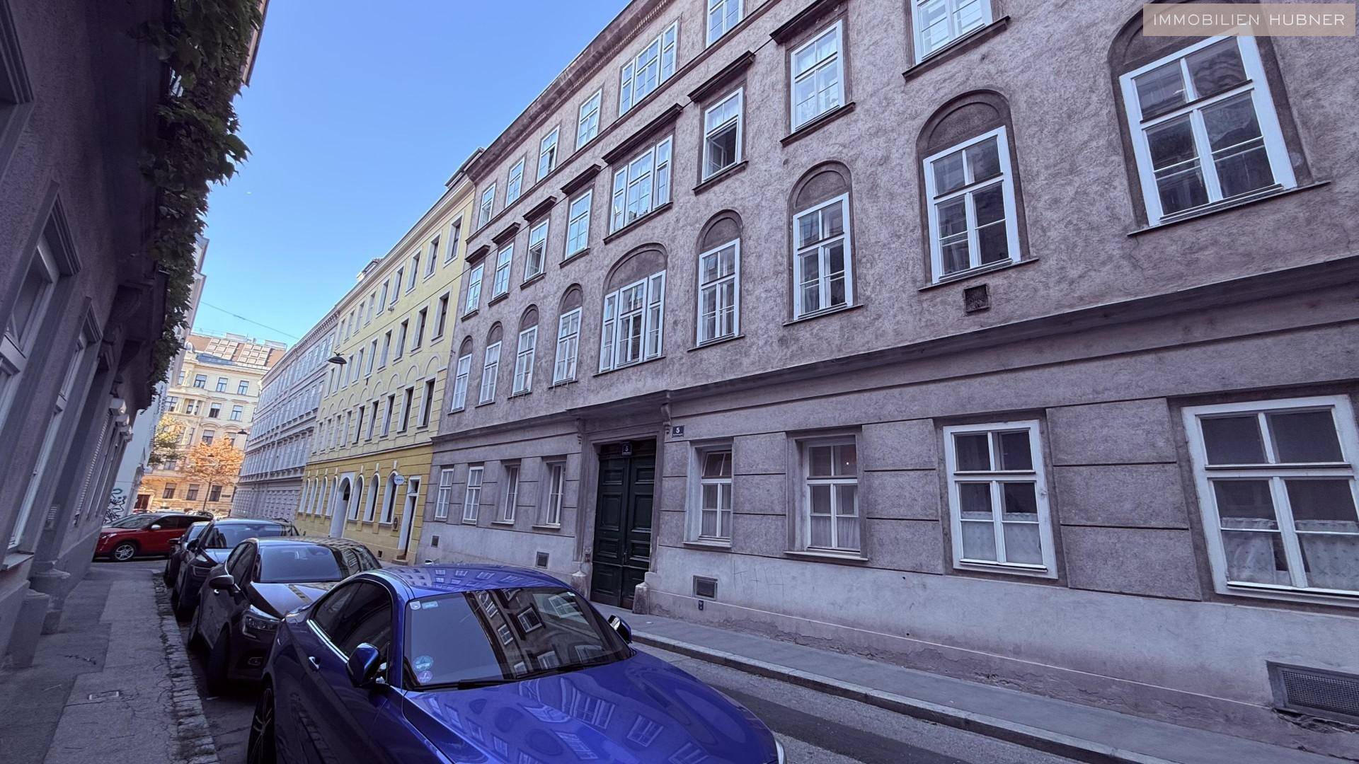 Ruhige Stadtstraße mit parkenden Autos und einer historischen Gebäudefassade unter blauem Himmel.