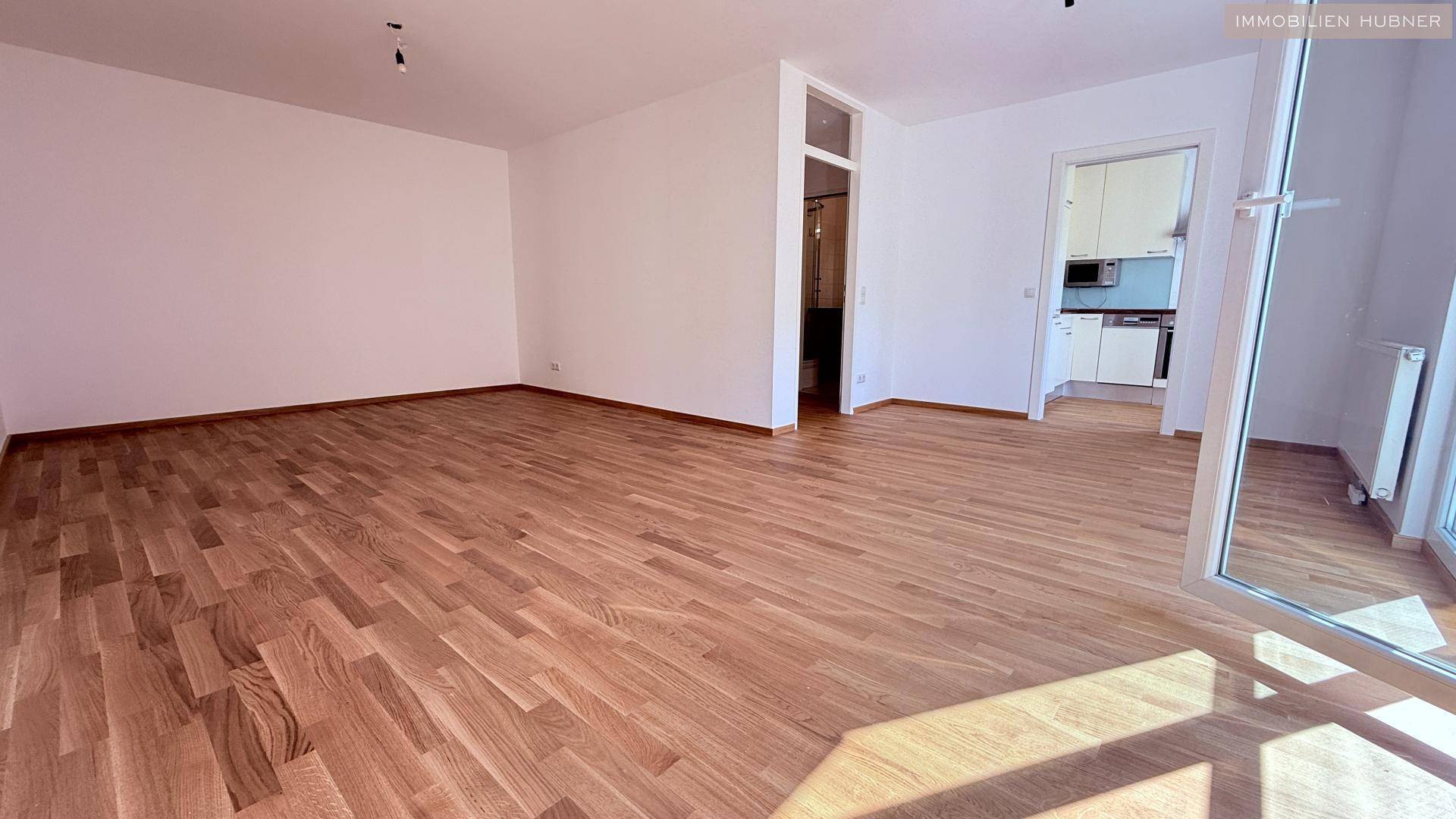 Geräumiges Zimmer mit hellem Holzparkettboden und Zugang zu einer modernen Küche.