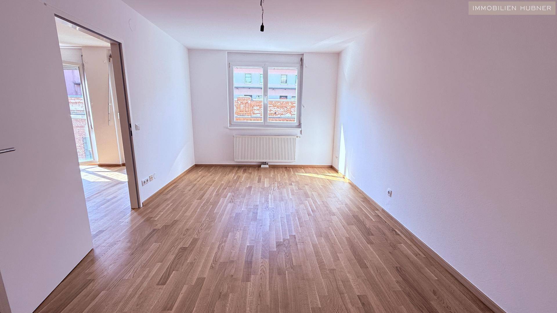 Leeres Zimmer mit hellem Holzparkettboden, weißen Wänden und einem Fenster mit Heizkörper.