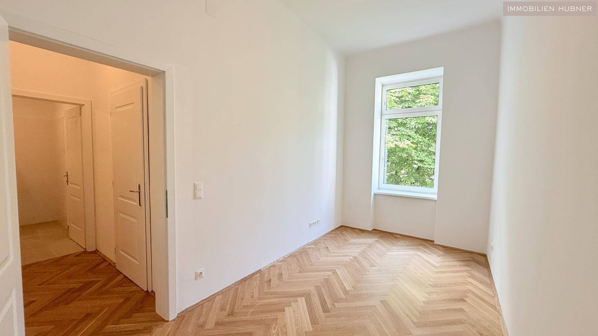 Helles Schlafzimmer mit Fischgrätparkettboden und großem Fenster mit Blick ins Grüne.