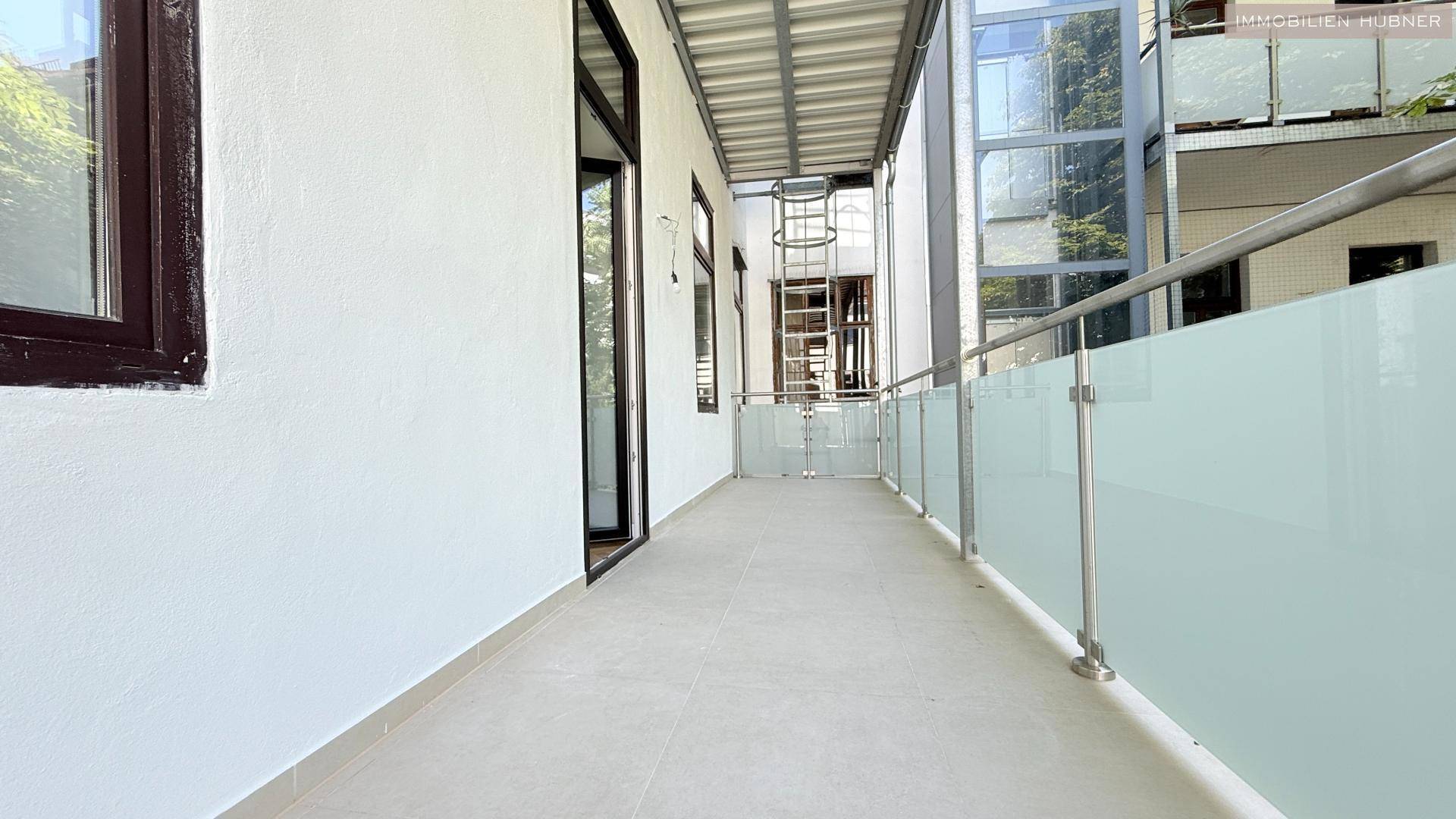 Geräumiger Balkon mit Glasgeländer und Blick auf den externen Aufzug des Gebäudes.