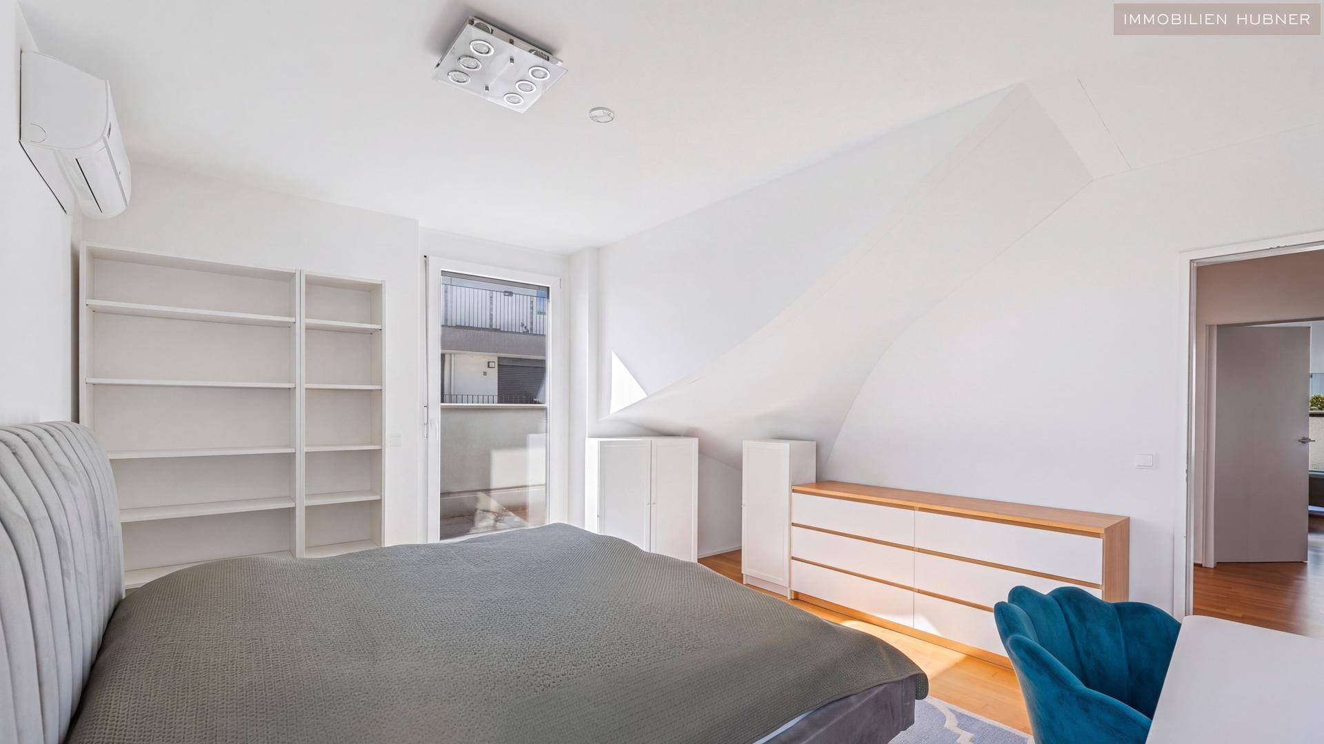 Helles Schlafzimmer mit Dachschräge, Einbauregalen und Zugang zu einem kleinen Balkon.