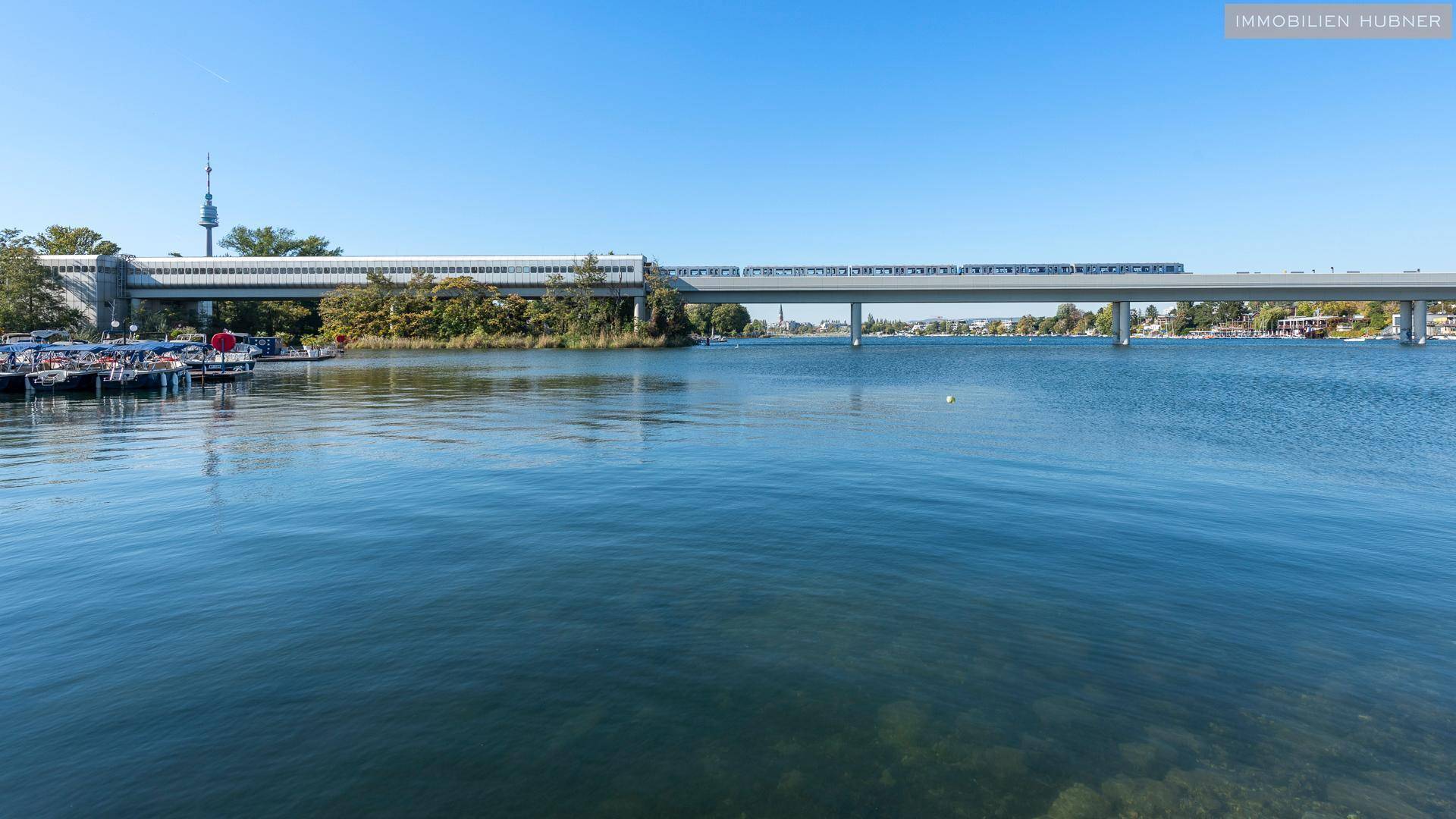 Ruhiger Blick auf den See mit einer modernen Brücke und einem Zug im Hintergrund.