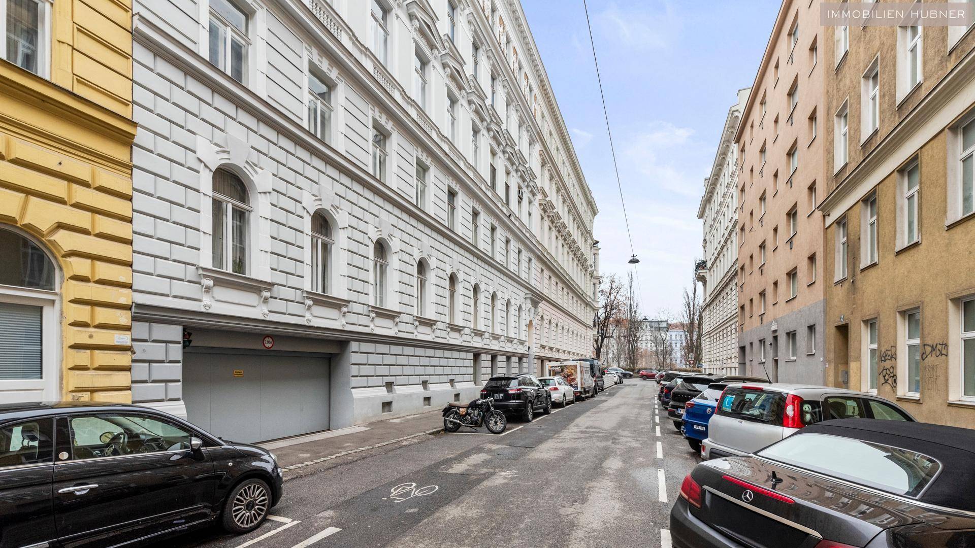 Historische Straßenansicht mit Altbaufassaden, geparkten Autos und einer Garageneinfahrt.