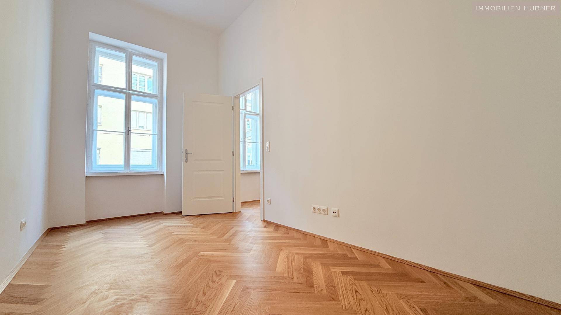 Geräumiges Zimmer mit zwei hohen Fenstern und klassischem Fischgrätparkett, ideal für vielseitige Nutzung.