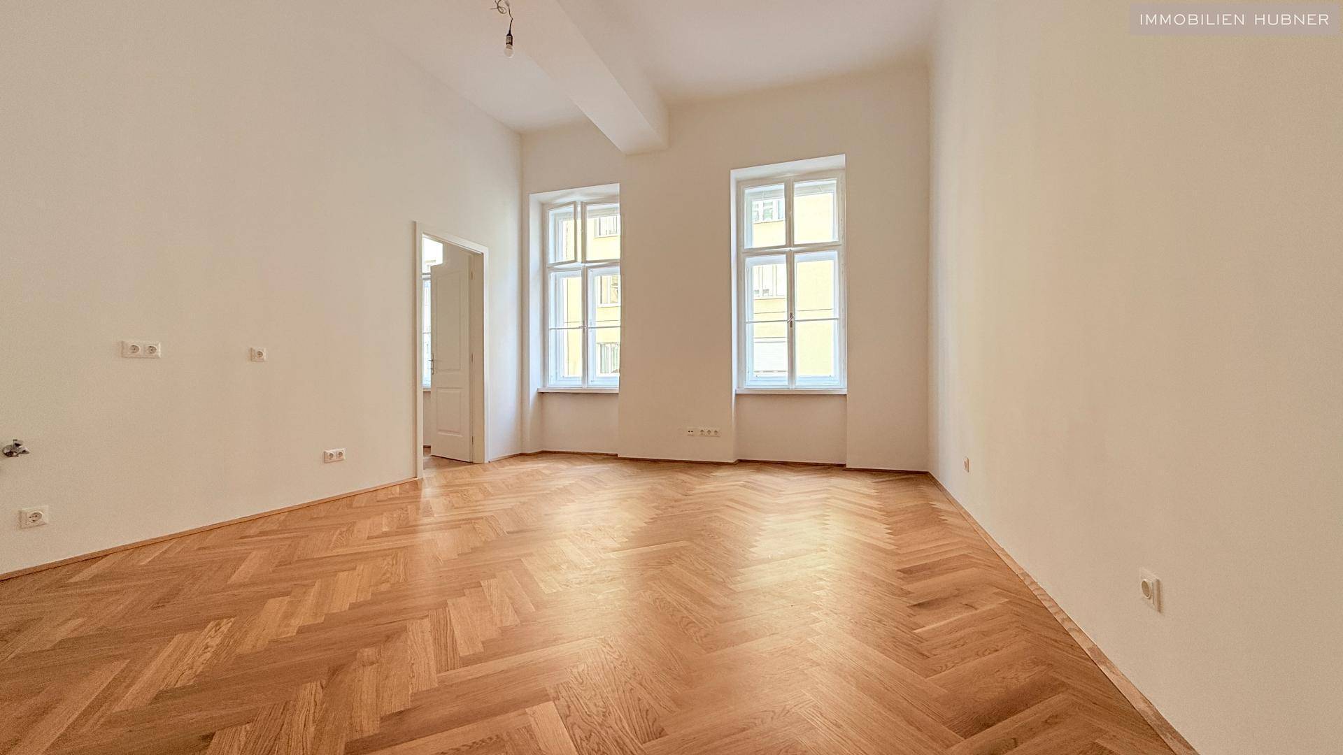 Lichtdurchflutetes Zimmer mit zwei großen Fenstern und elegantem Fischgrätparkett.