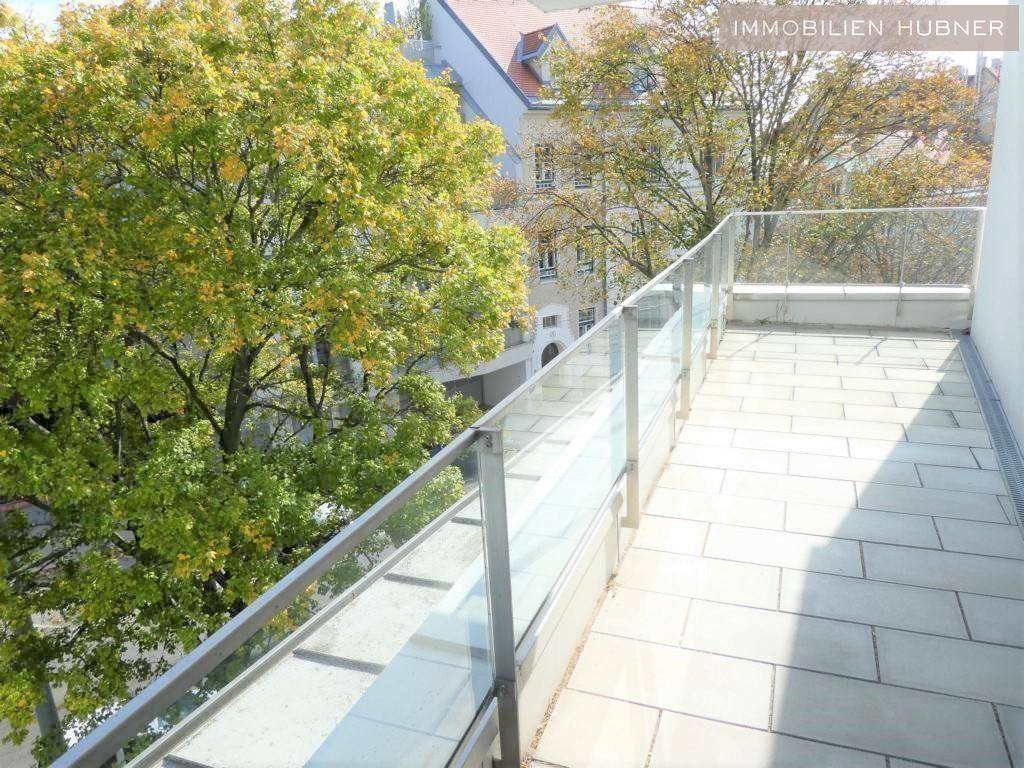 Langer Balkon mit Glasgeländer und Blick auf grüne Bäume und umliegende Gebäude.
