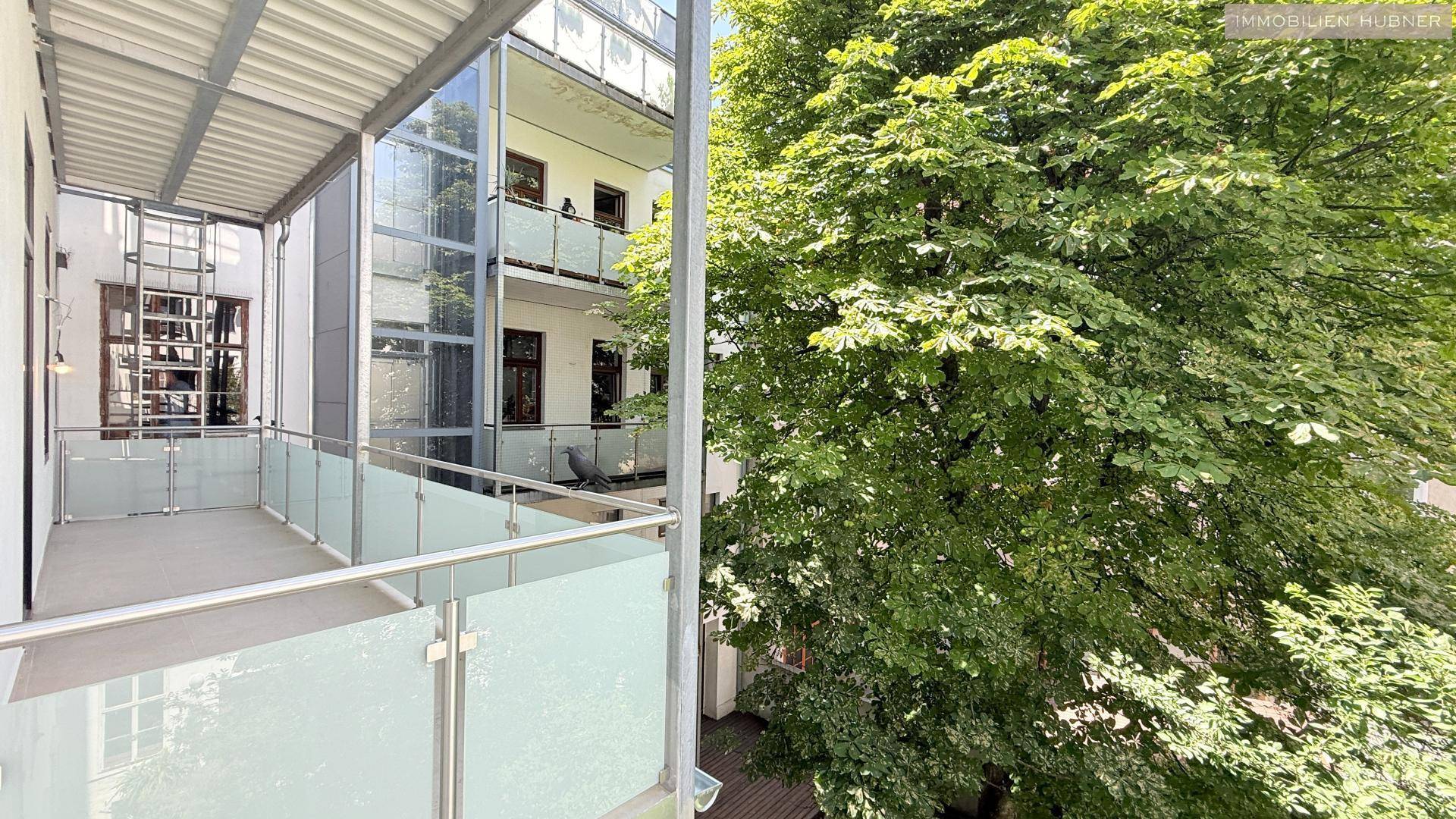 Heller Balkon mit Glasgeländer und üppigem Grünblick, angrenzend an ein Gebäude mit Aufzug.