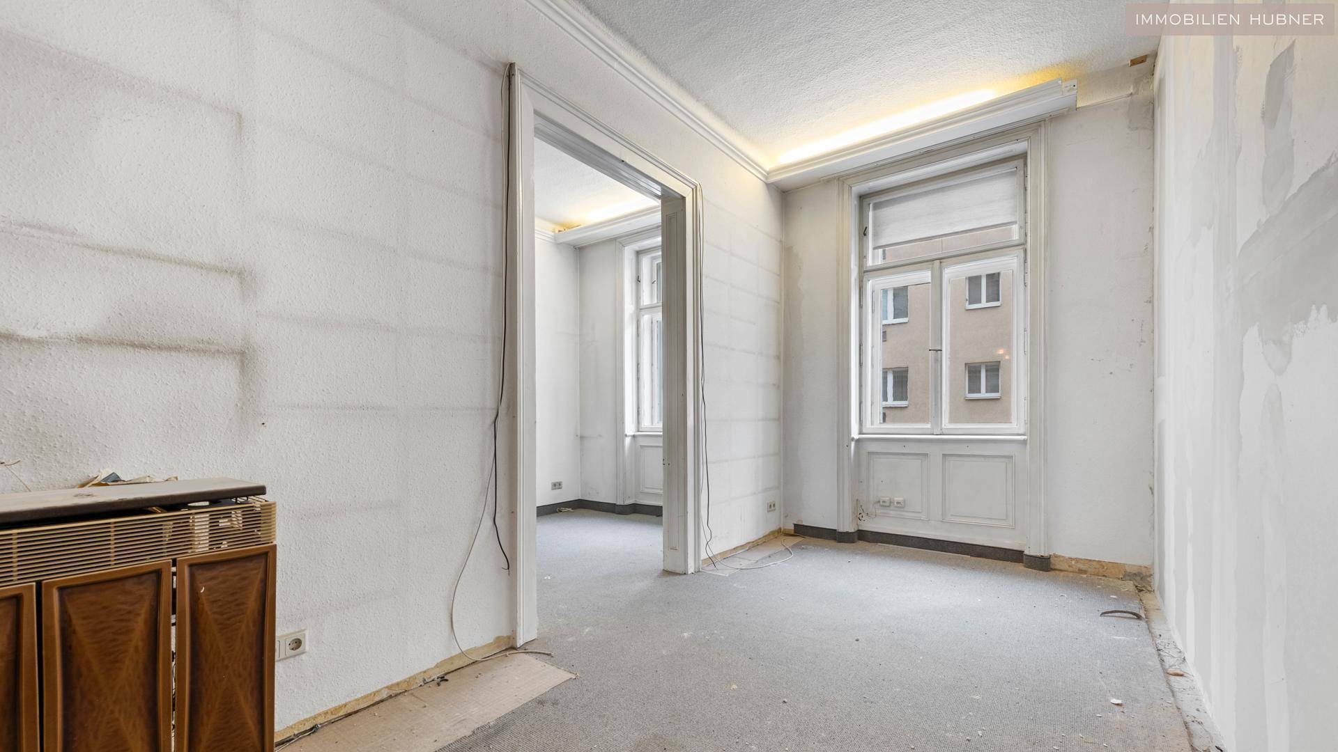 Renovierungsbedürftiger Raum mit hohen Decken, großen Fenstern und Blick auf die Nachbargebäude.