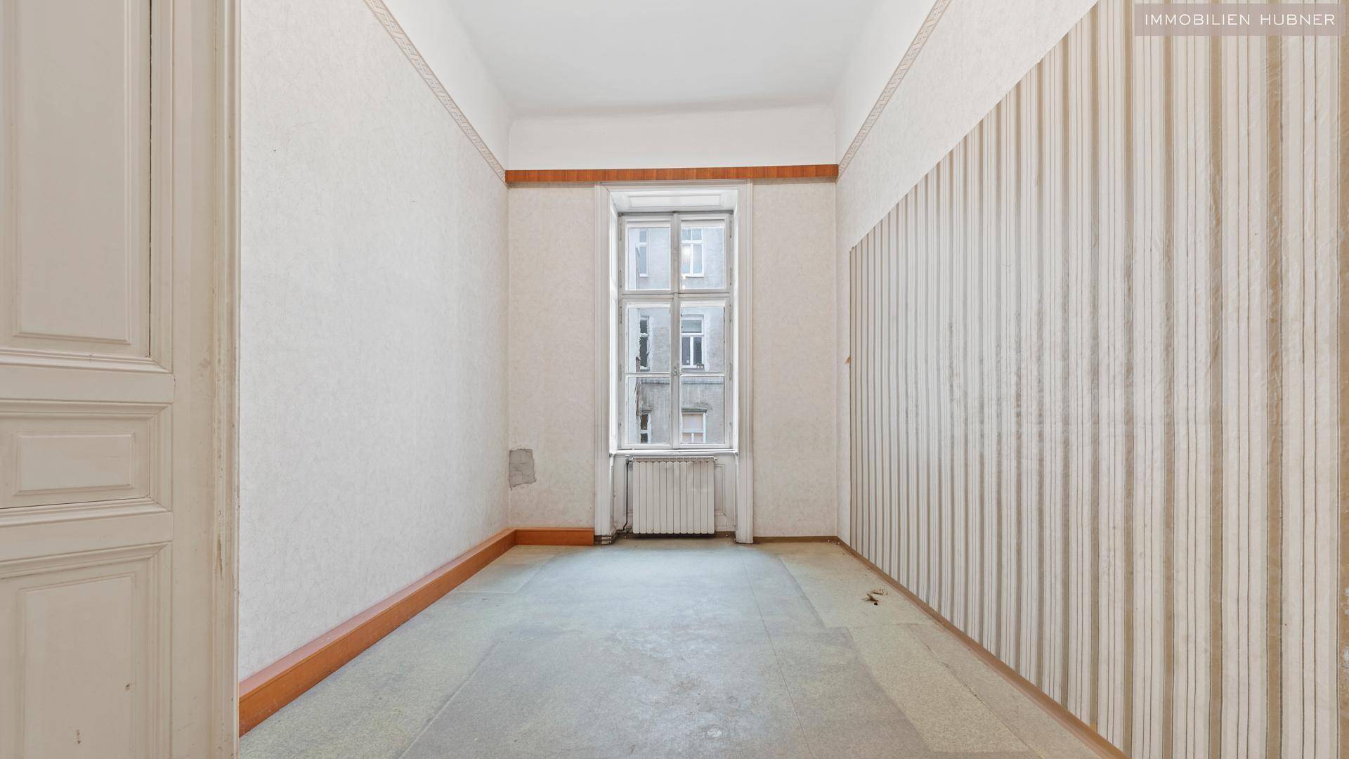 Leerer Raum mit Fenster, Heizkörper und gestreifter Tapete, der auf Renovierung wartet.